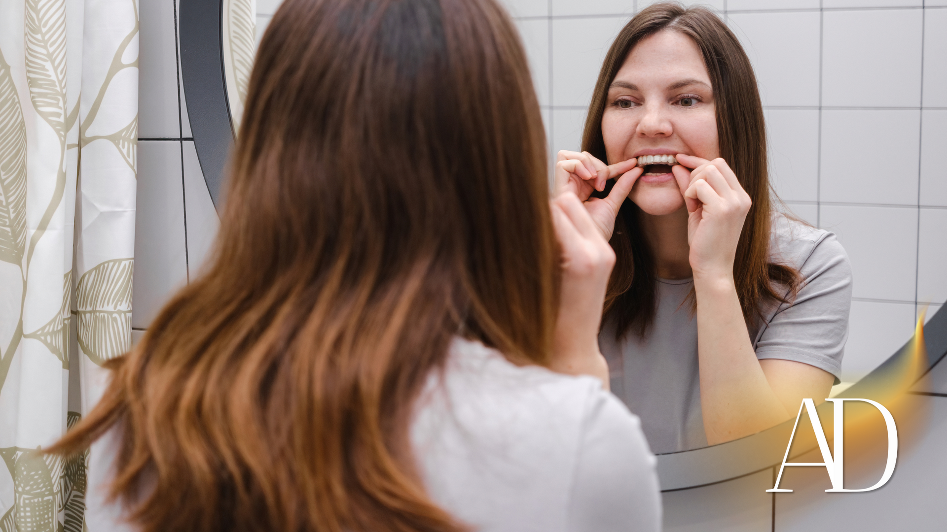 Woman flossing teeth in bathroom mirror, with white tile background and shower curtain.