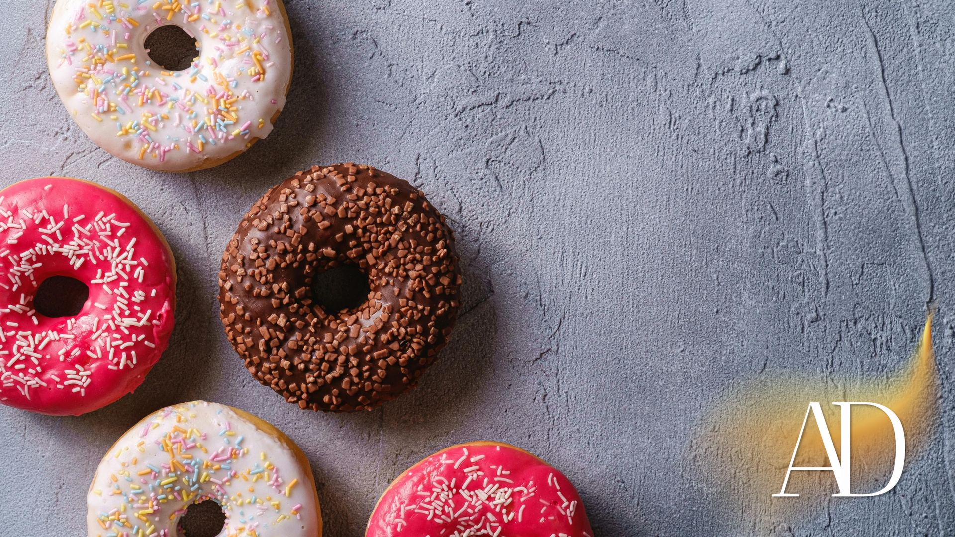 Assorted frosted donuts on a gray surface, including pink, white, and chocolate with sprinkles.