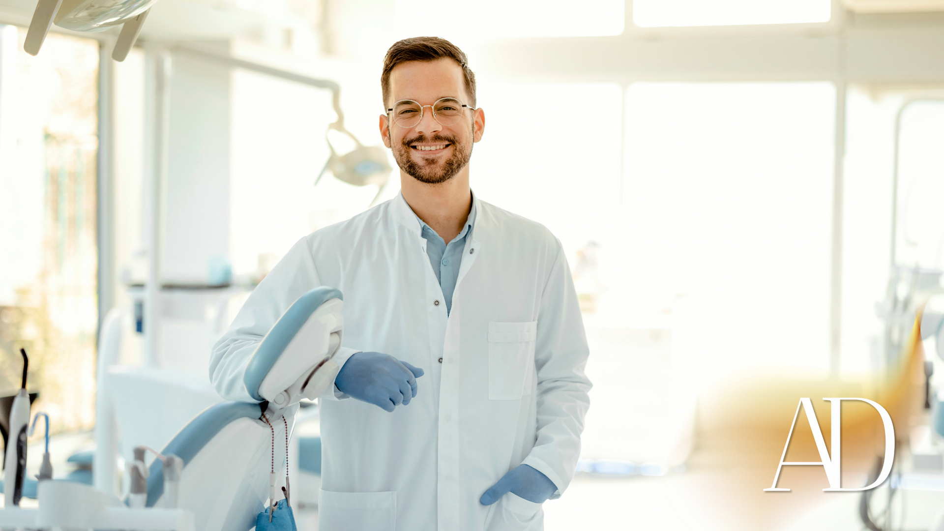 Dentist in white coat and blue gloves standing beside dental equipment in a bright clinic