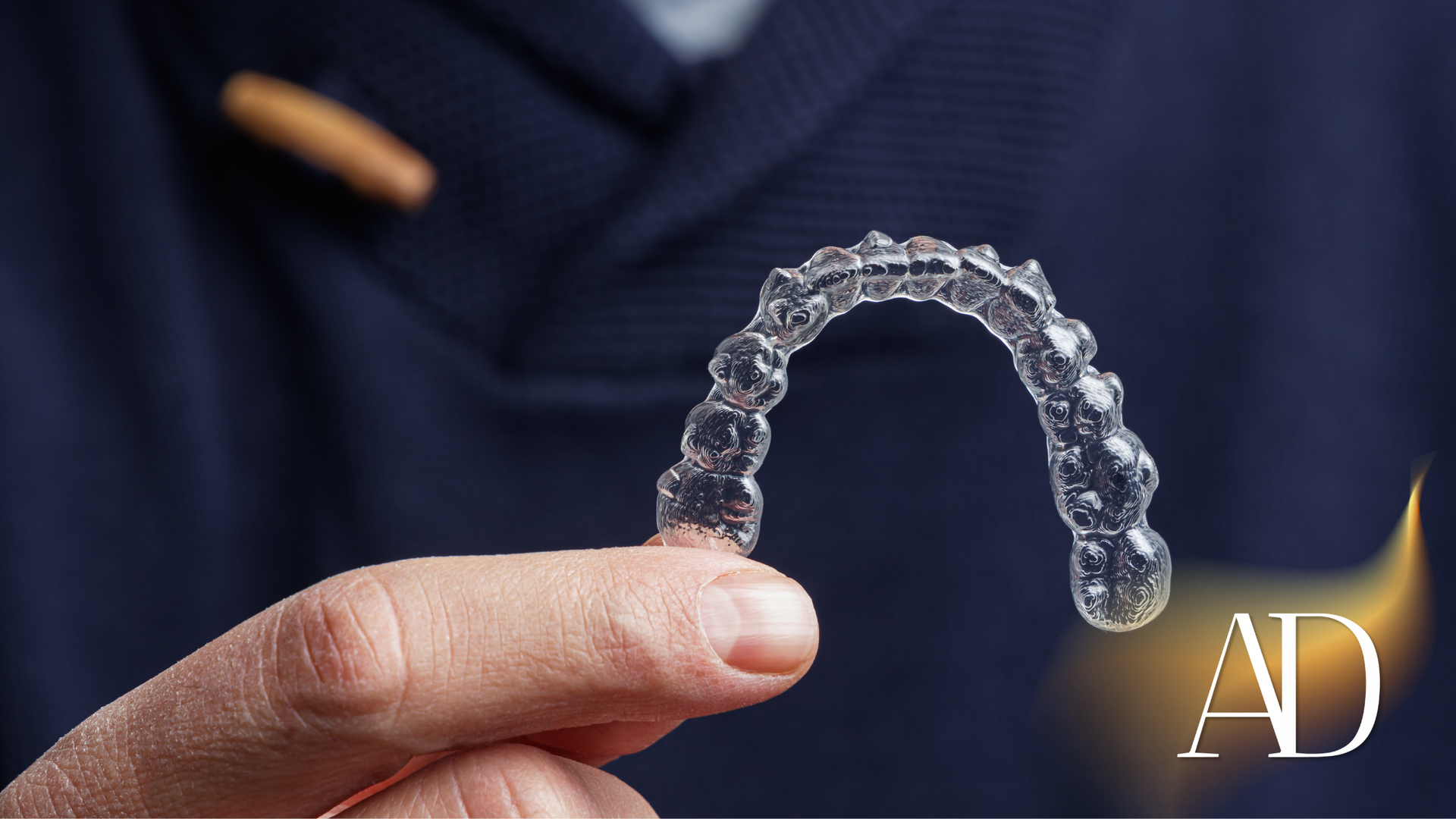 Hand holding a clear dental aligner, with a blurred person in a dark shirt in the background