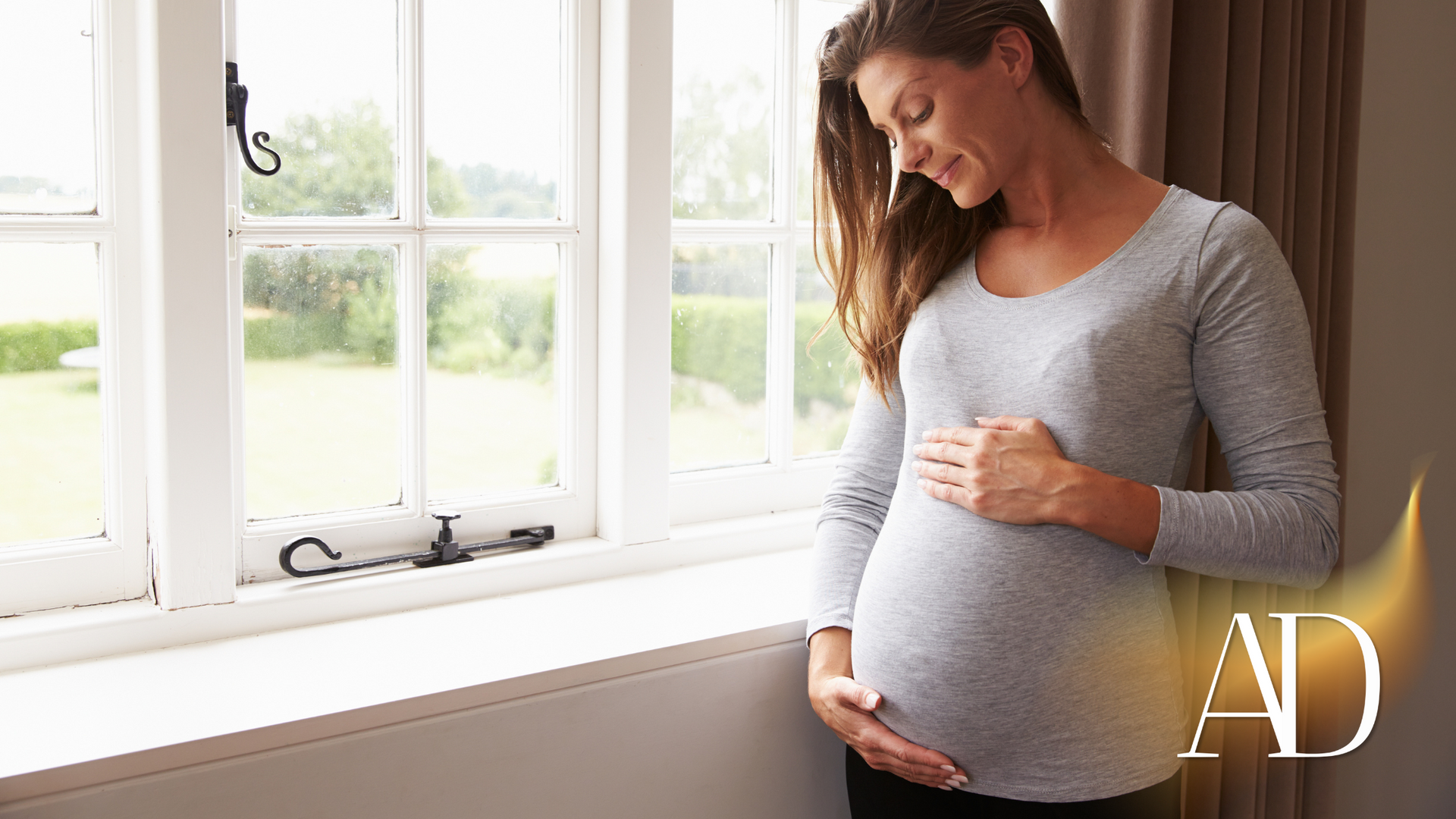 Pregnant woman in a gray dress gently holding her belly by a window