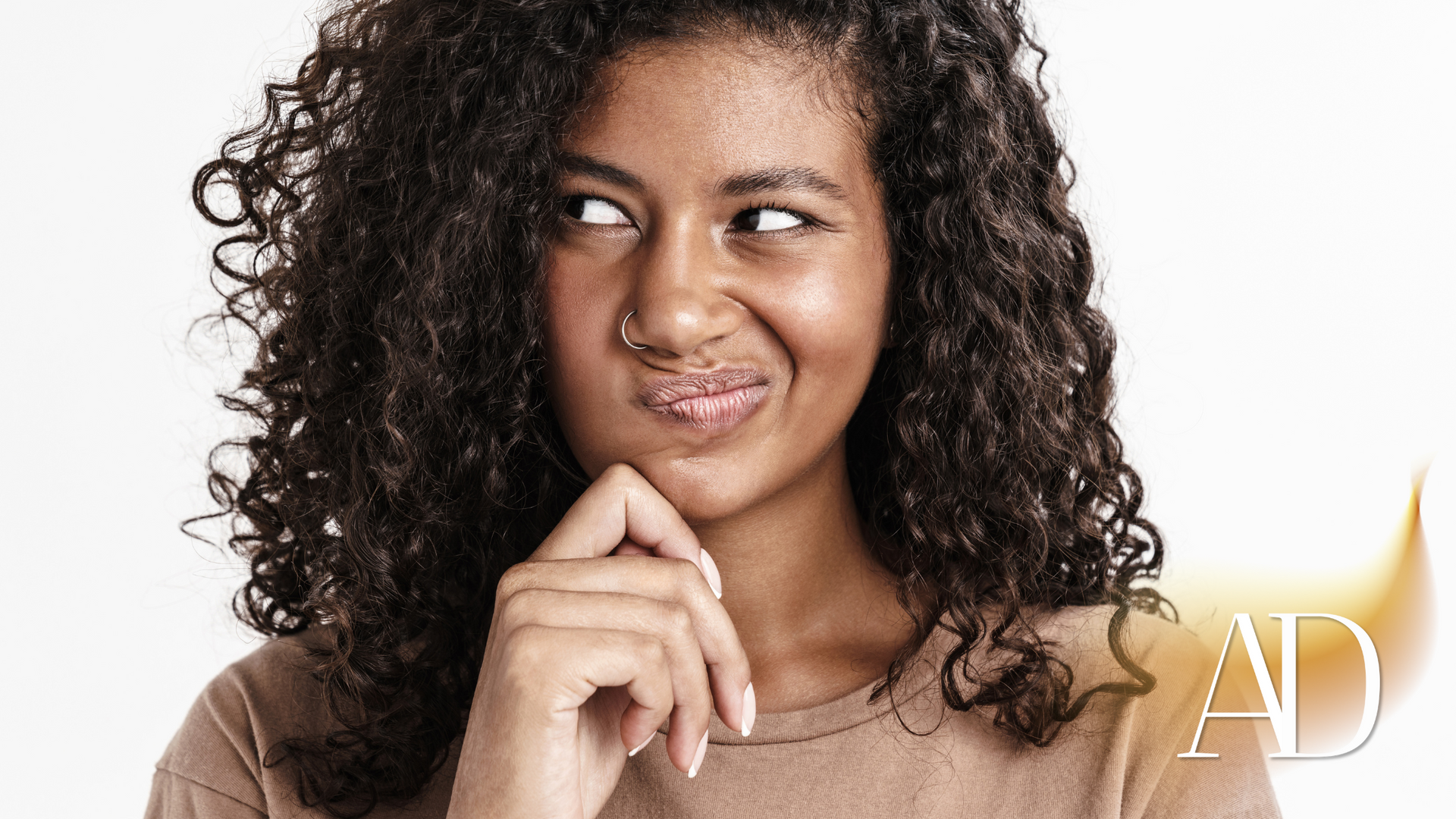 Curly-haired woman in a beige top, smirking and looking to the side with her hand on her chin.