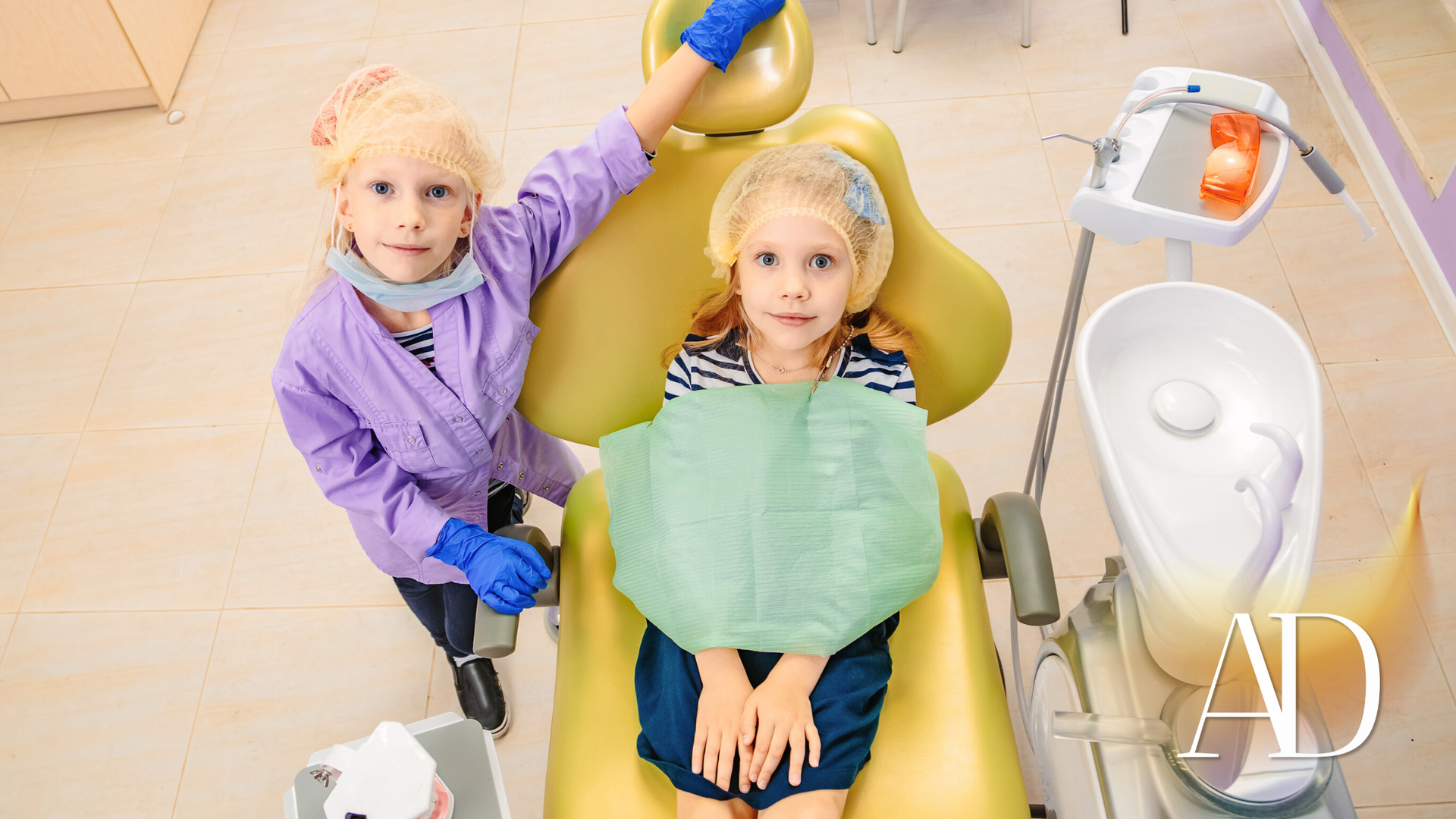 Child in a dental chair with green bib while a child in a purple coat stands beside them in a dental clinic