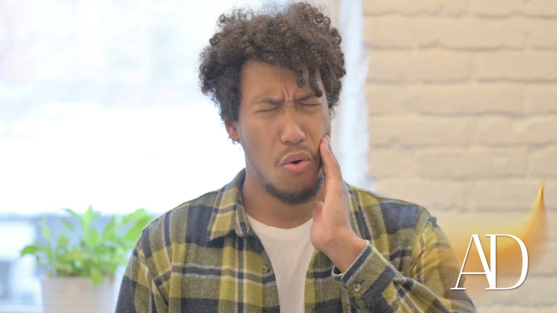 Man grimacing and holding his cheek, wearing a yellow plaid shirt indoors near a bright window.
