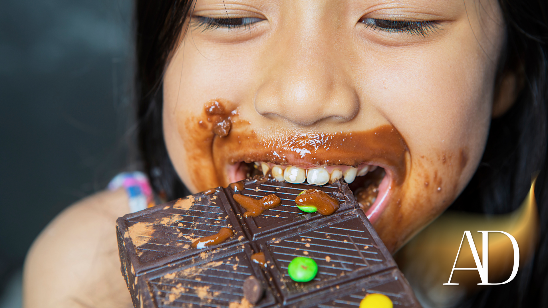 Child biting a chocolate bar, face smeared with melted chocolate and colorful candies scattered on top