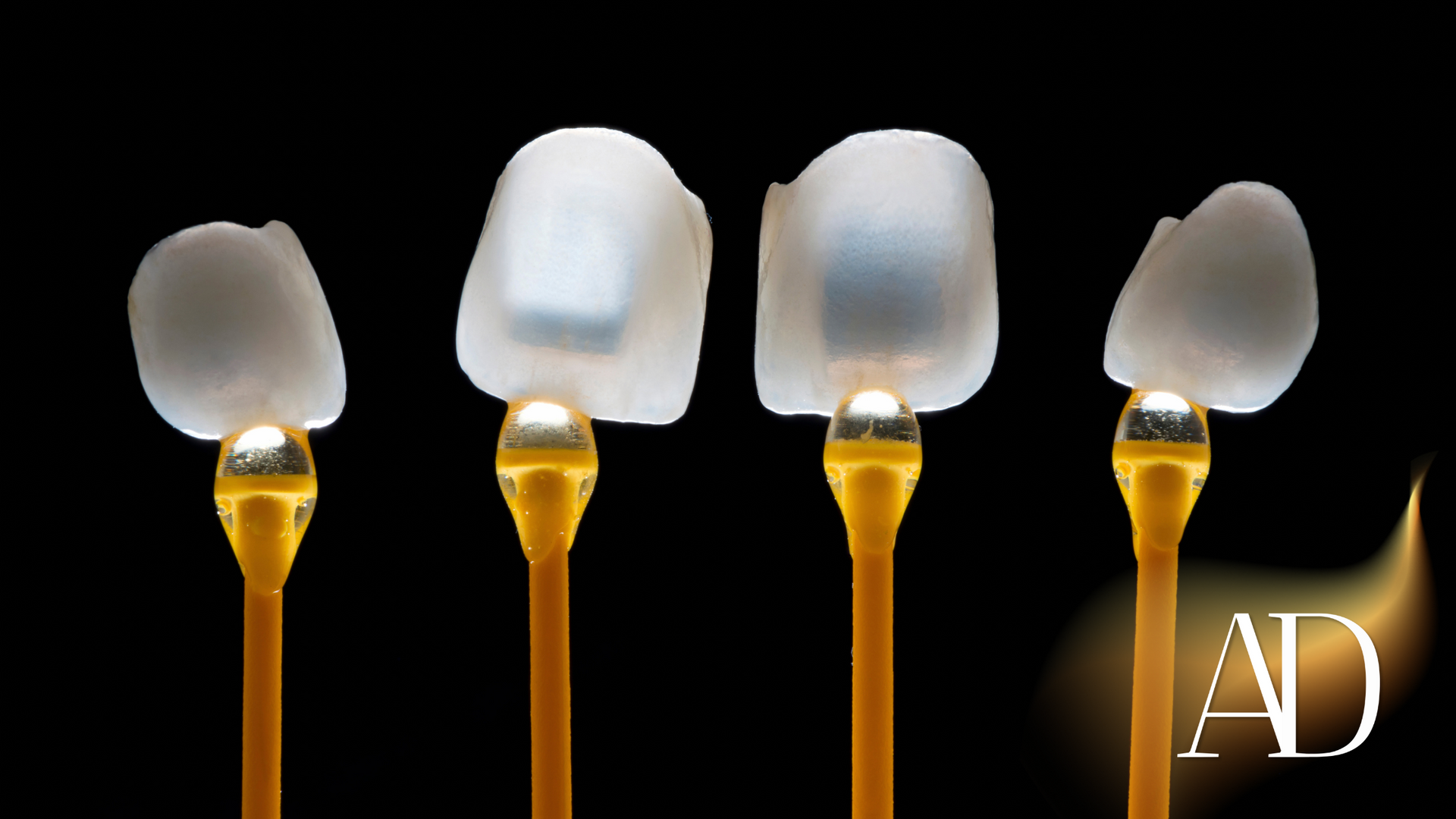 Four cotton swabs with translucent white tips on gold stems against a black background, one marked “AD”
