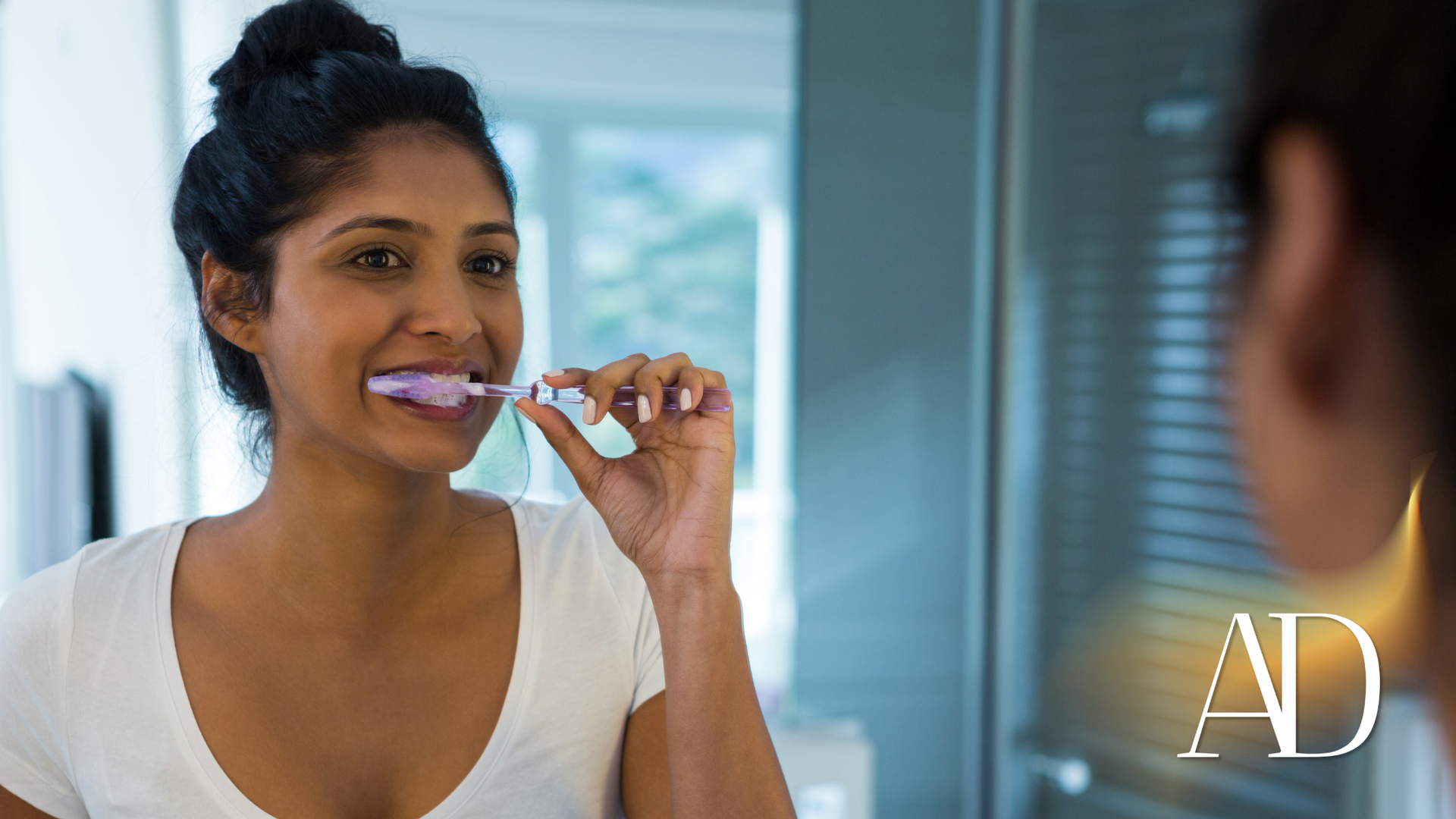 Woman brushing her teeth in a bathroom mirror, wearing a white shirt.