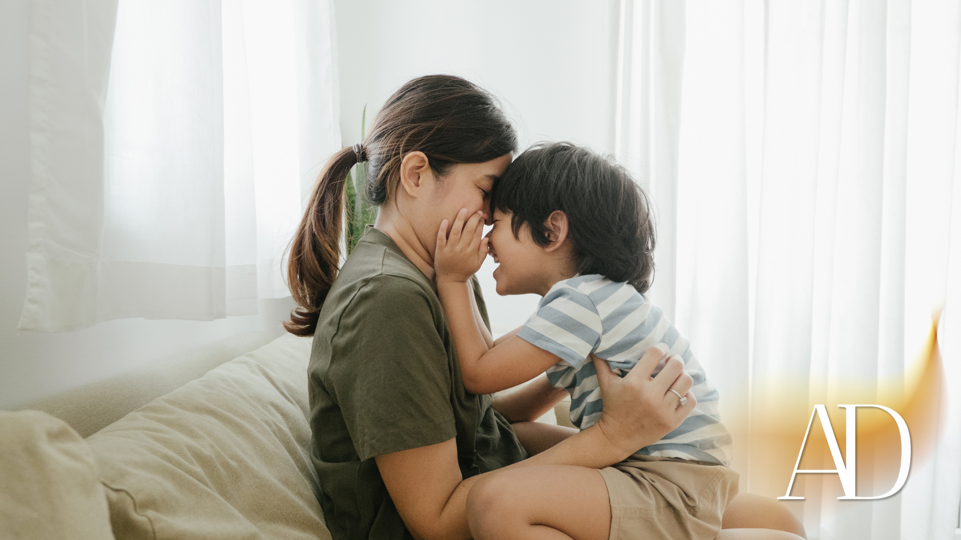 Parent and child sitting on a couch, touching foreheads in a bright room with sheer white curtains.