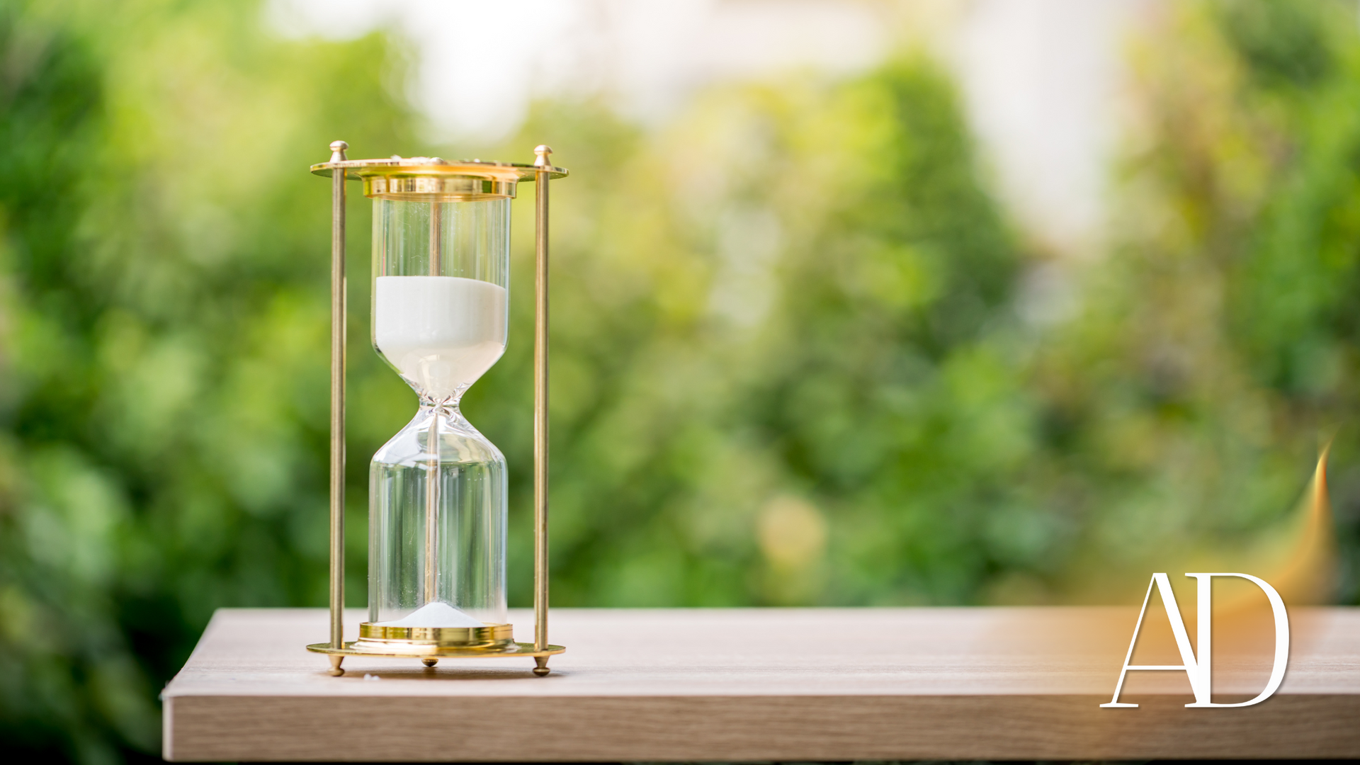 White hourglass in a gold frame on a wooden table, with a blurred green outdoor background.