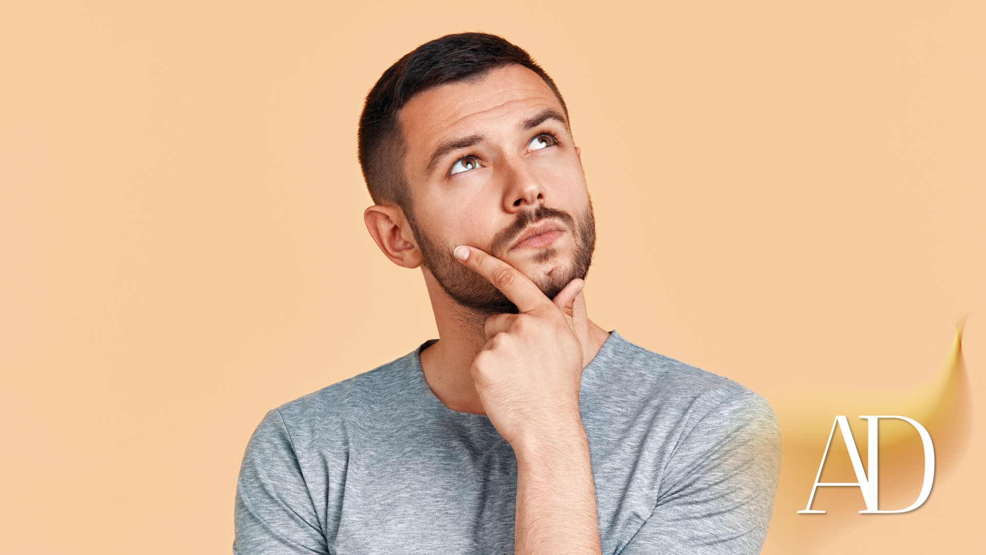 Man in gray shirt resting chin on hand and looking upward against a peach background