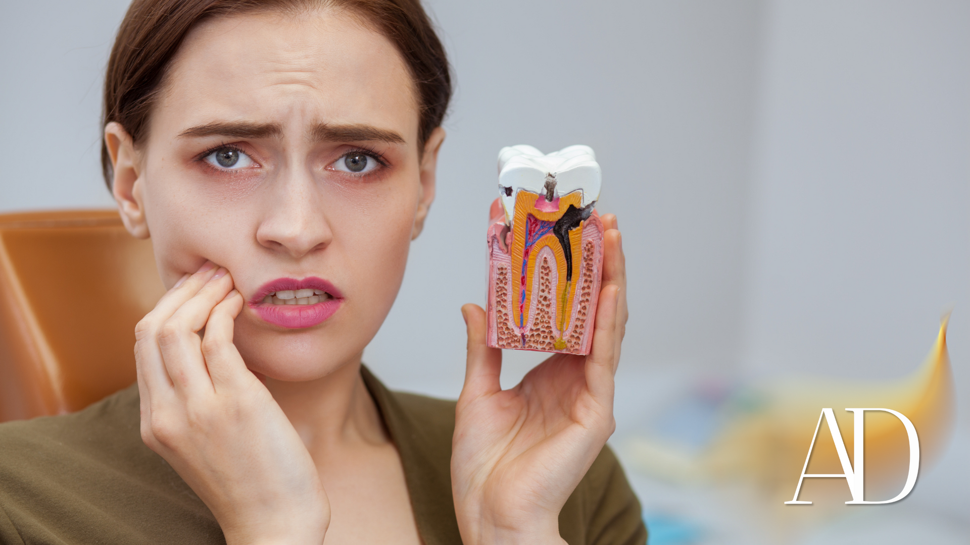 Woman holding a model tooth, touching her cheek with a pained expression, suggesting toothache.