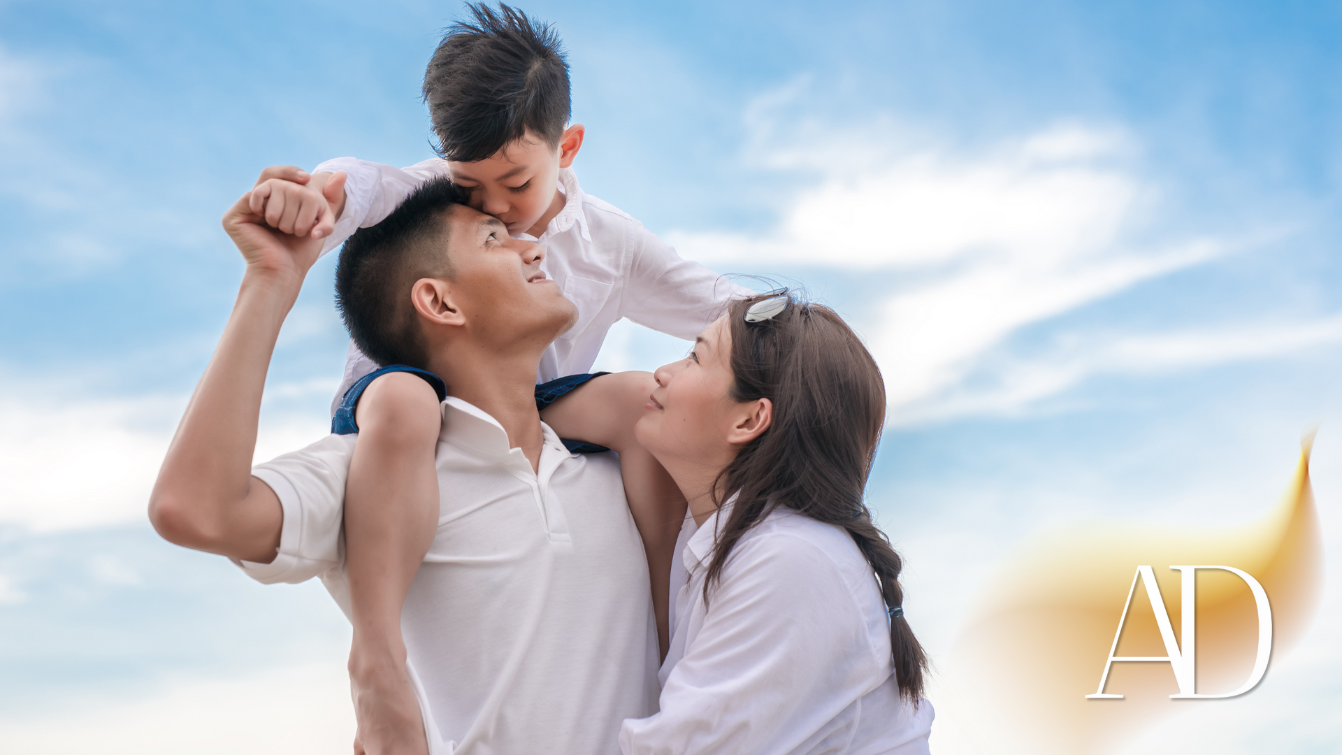 Family playing together outdoors under a blue sky, parents lifting a child on their shoulders while smiling