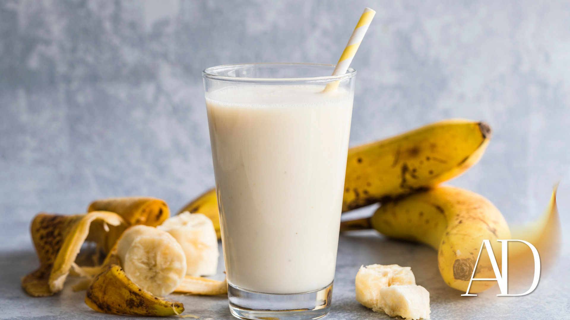 Glass of banana smoothie with a striped straw, surrounded by bananas and banana peels on a gray surface.