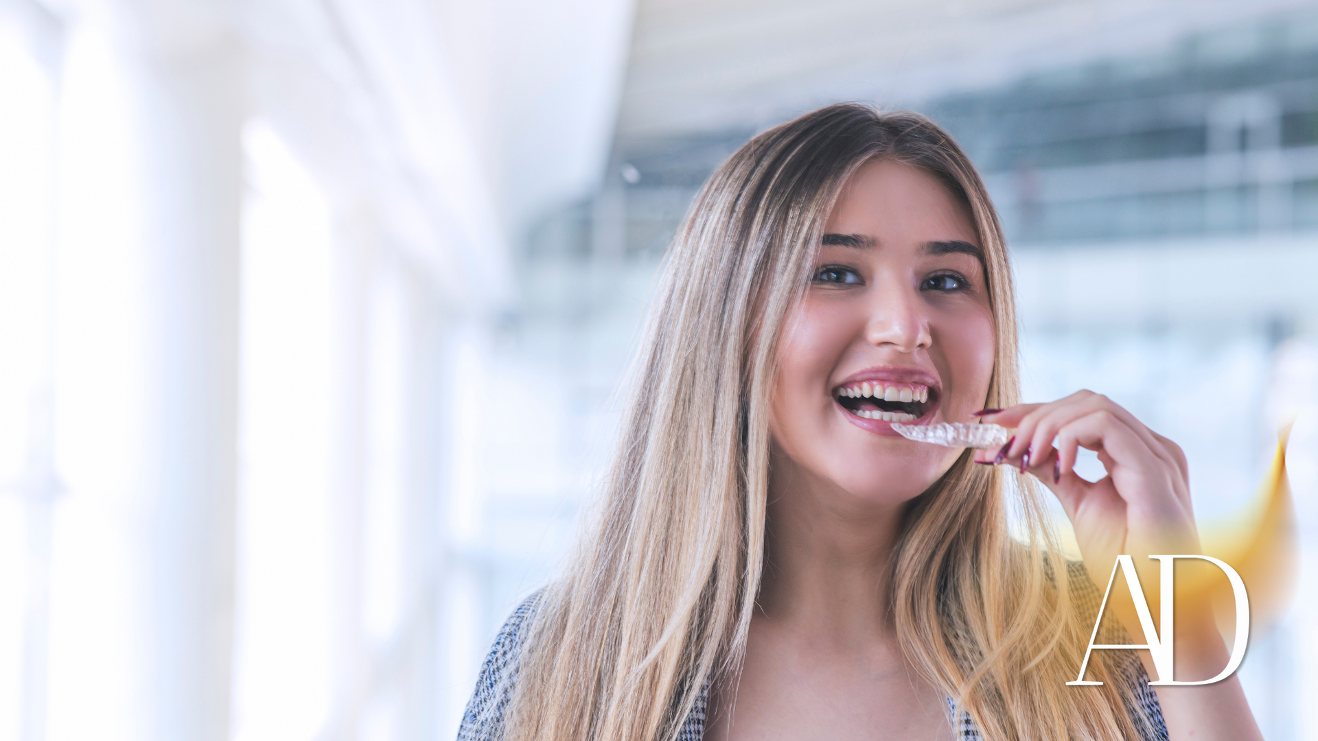 Smiling woman brushing her teeth in a bright bathroom, with a white towel over her shoulder.