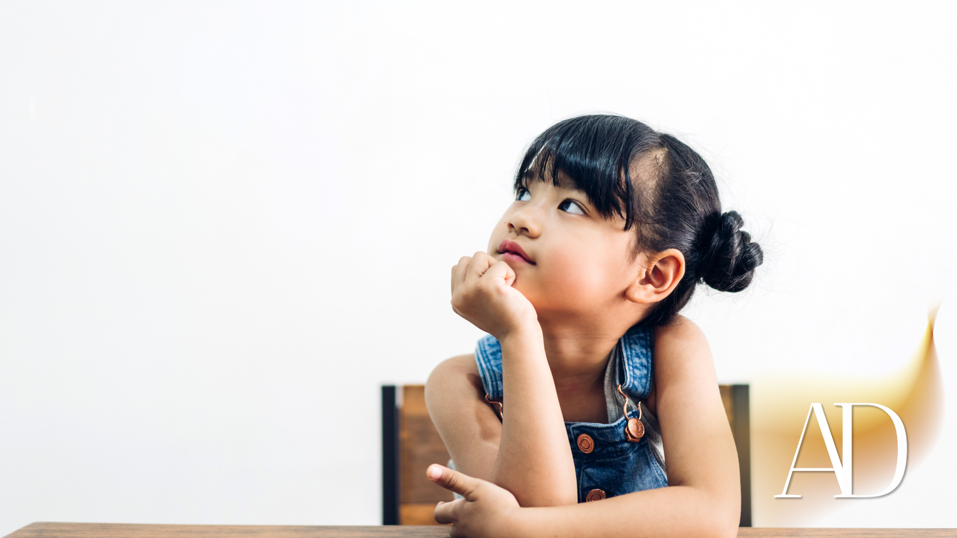 Young child with braided hair resting chin on hand at a table, looking up against a white background.