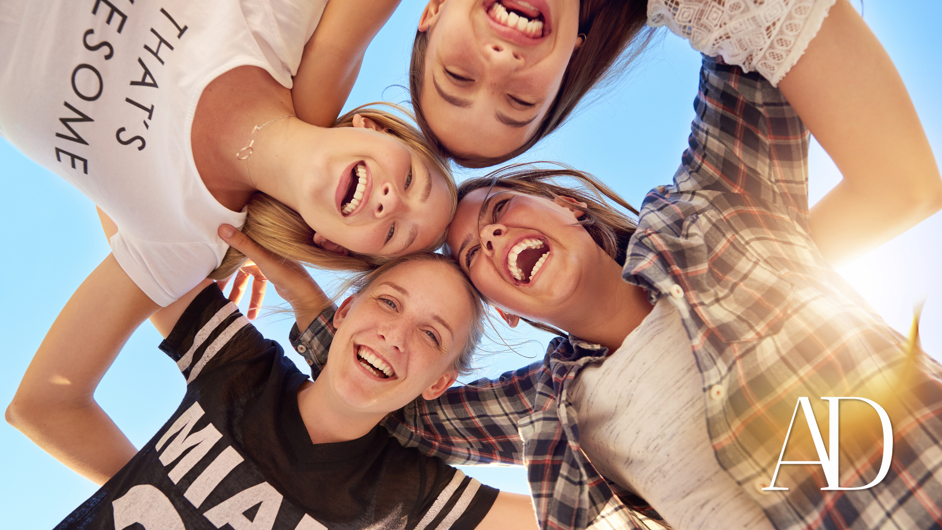Smiling group of friends huddle in a circle against a blue sky, viewed from below.