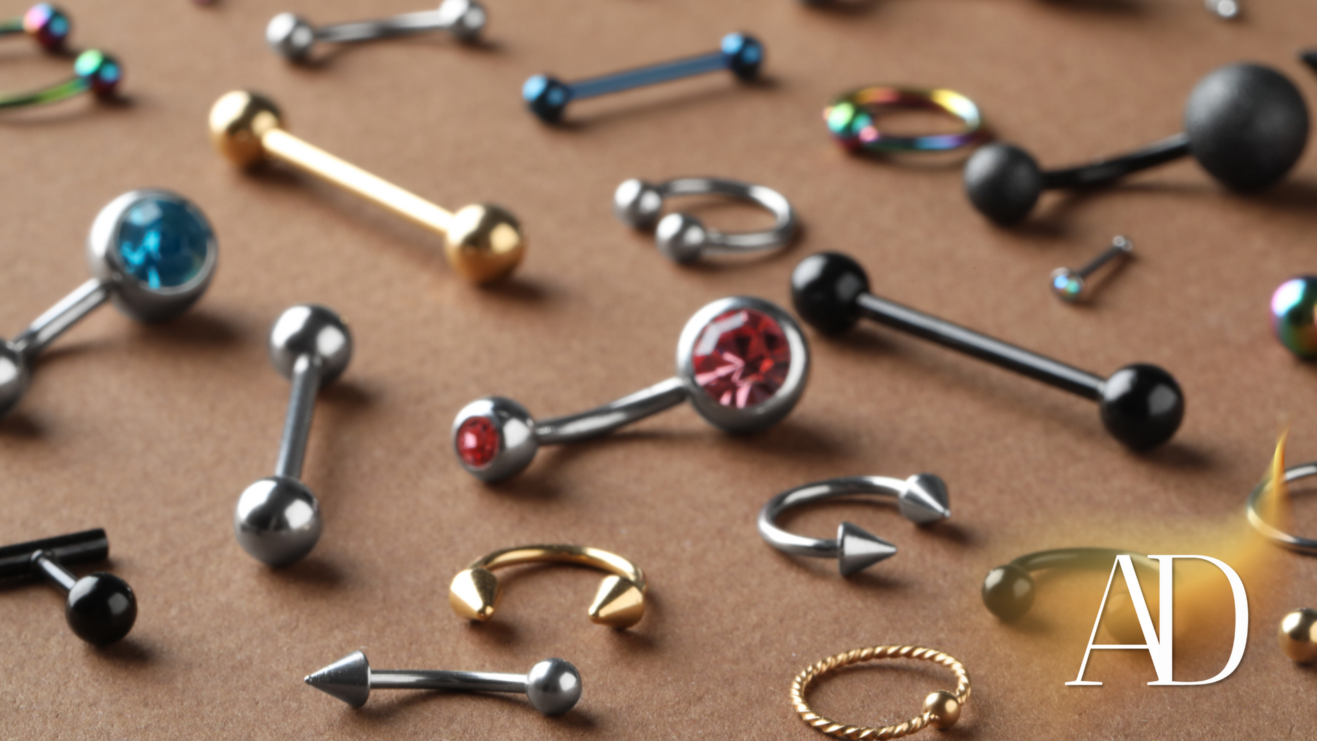 Assorted body jewelry studs and rings scattered on a tan surface, including blue, black, silver, and gold pieces.