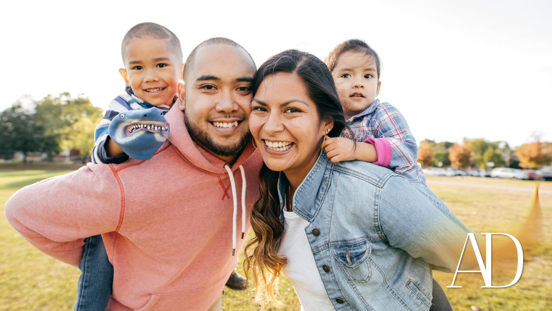 Smiling family of four outdoors in a grassy park, with a child on each parent’s shoulders.
