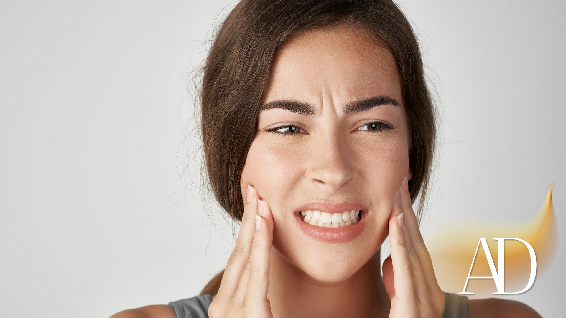 Woman grimacing and holding her cheek, suggesting tooth pain, against a light background.