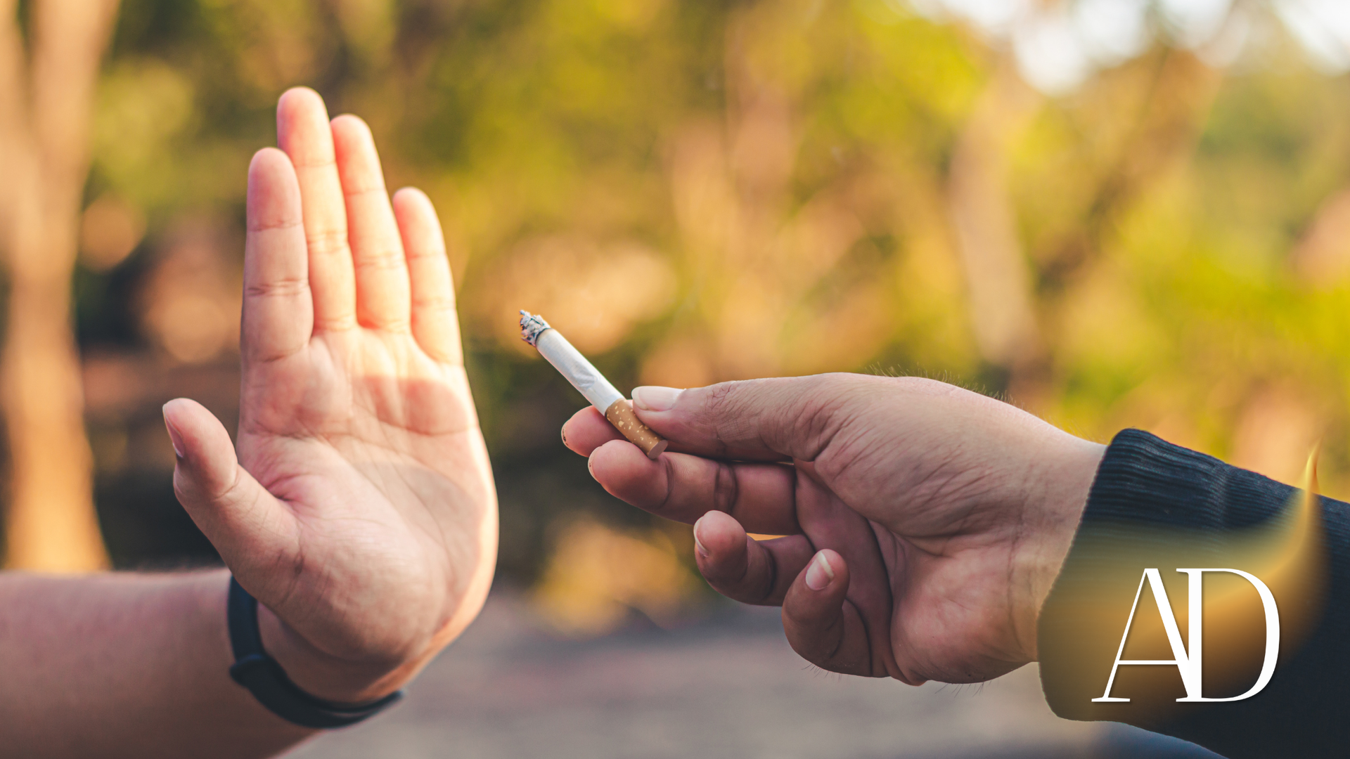 A hand holds a cigarette toward a raised palm in a blurred outdoor setting.