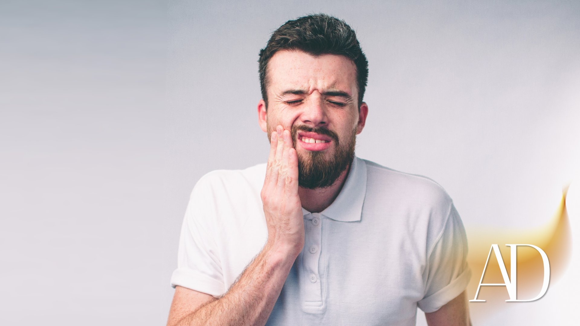 Man in a white shirt holding his cheek in discomfort against a light gray background