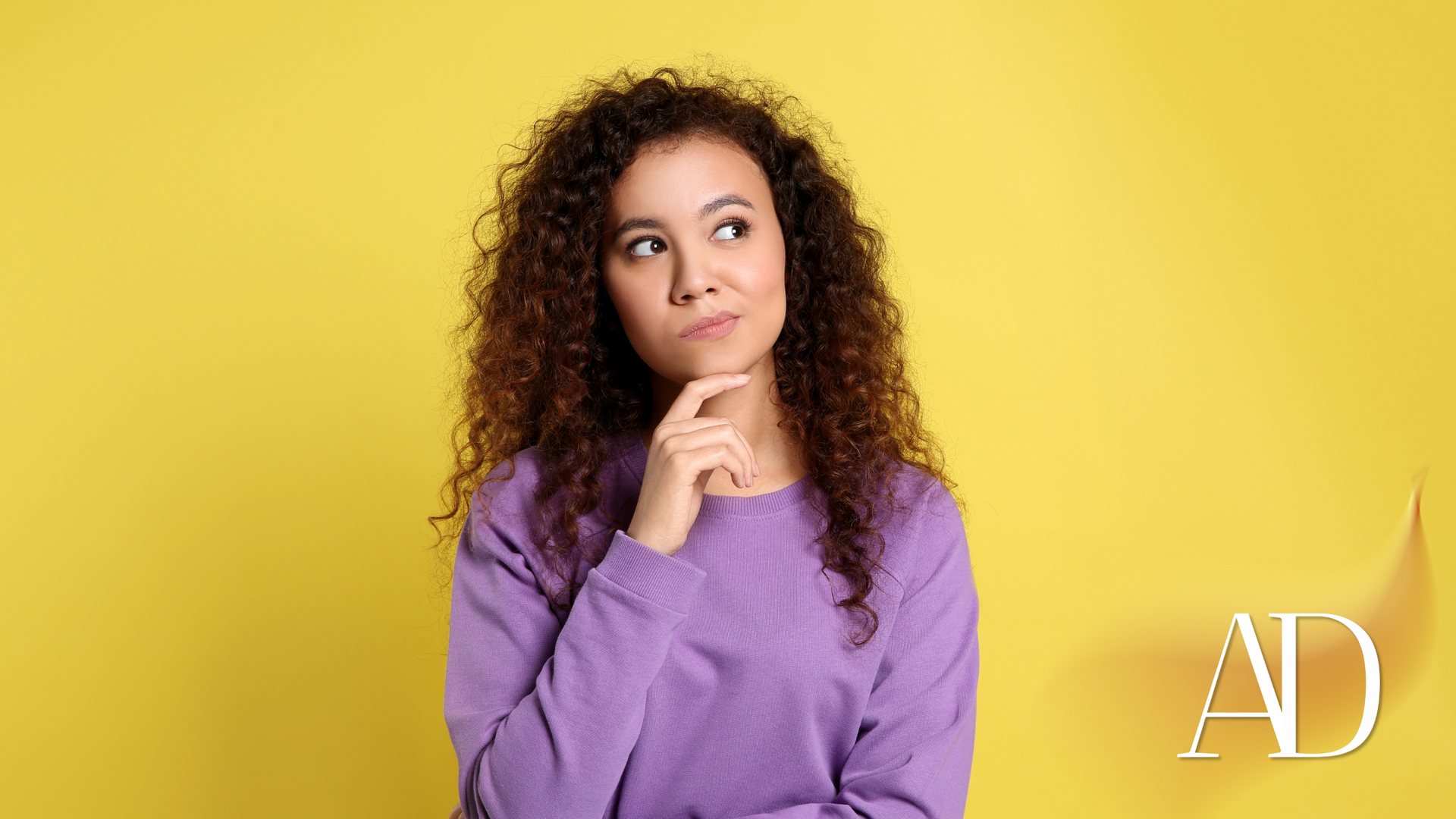 Curly-haired woman in a purple sweater looking up thoughtfully against a yellow background