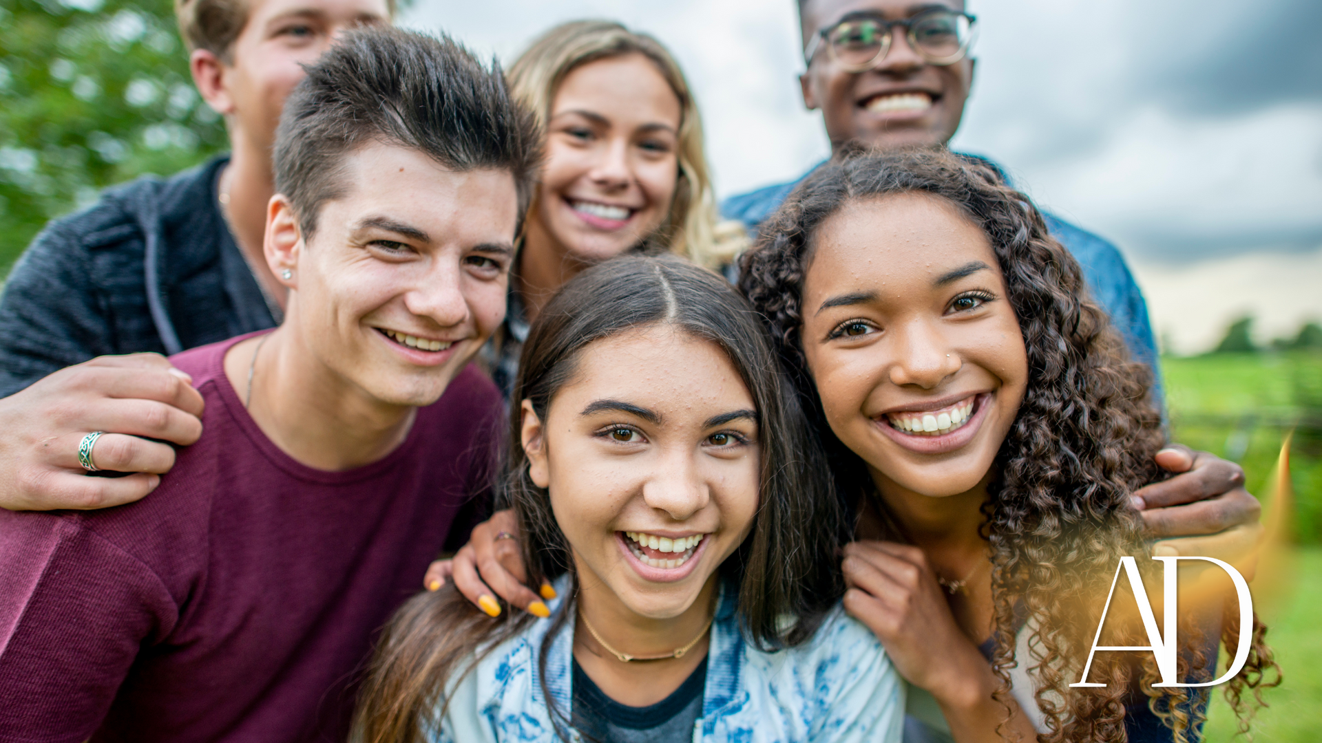 Smiling group of five friends posing outdoors in a grassy field under a cloudy sky