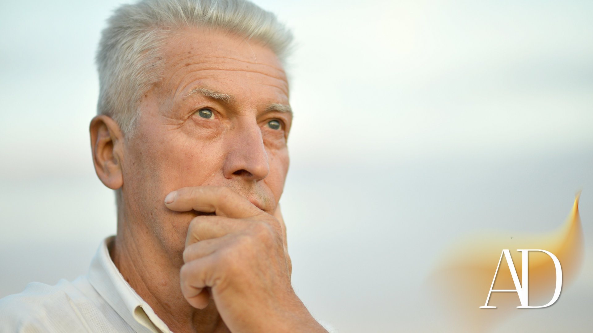 Thoughtful man with gray hair resting his hand on his chin against a bright sky, with an “AD” logo in the corner
