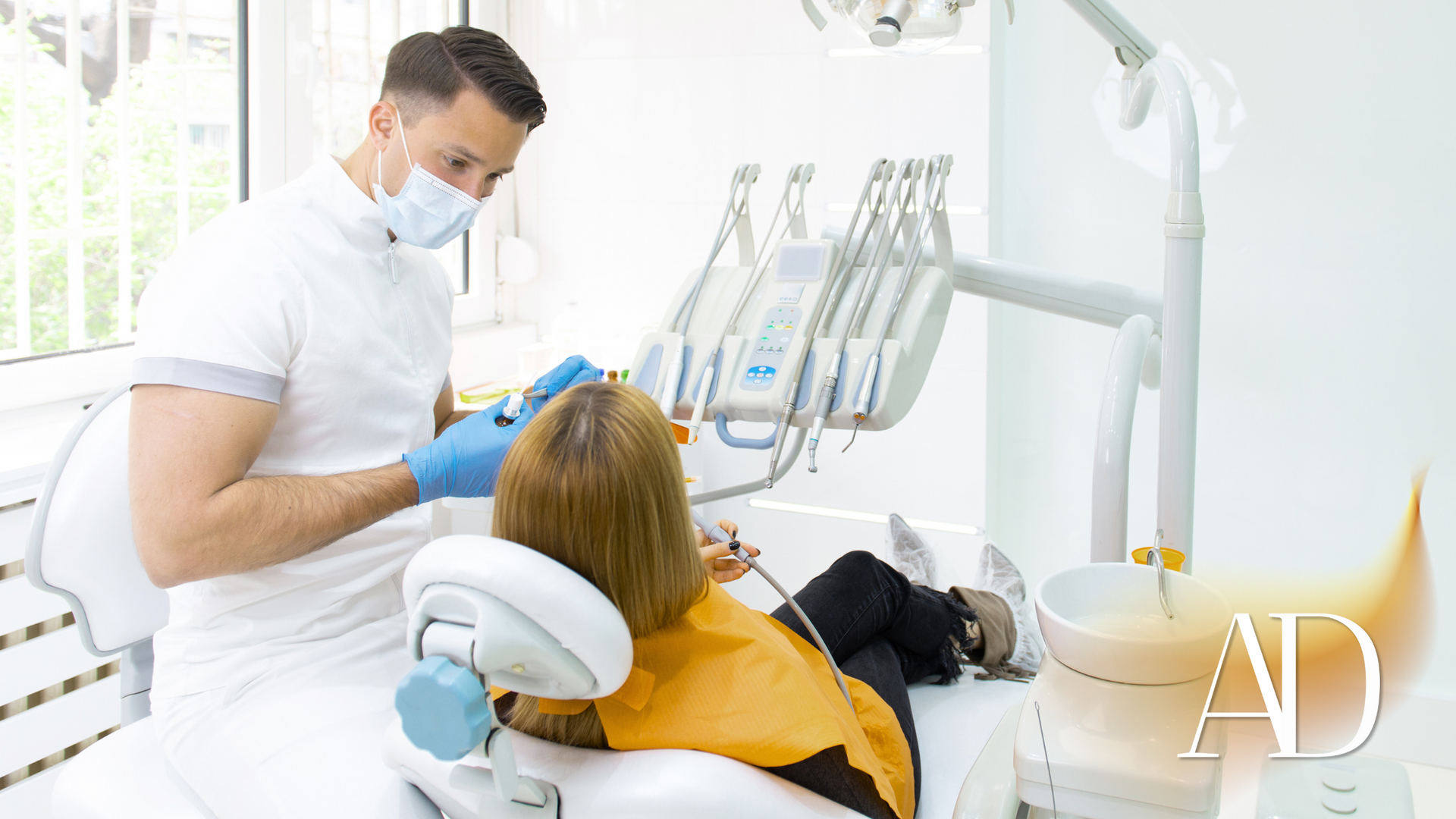 Dentist wearing a mask examines a patient reclined in a dental chair in a bright clinic