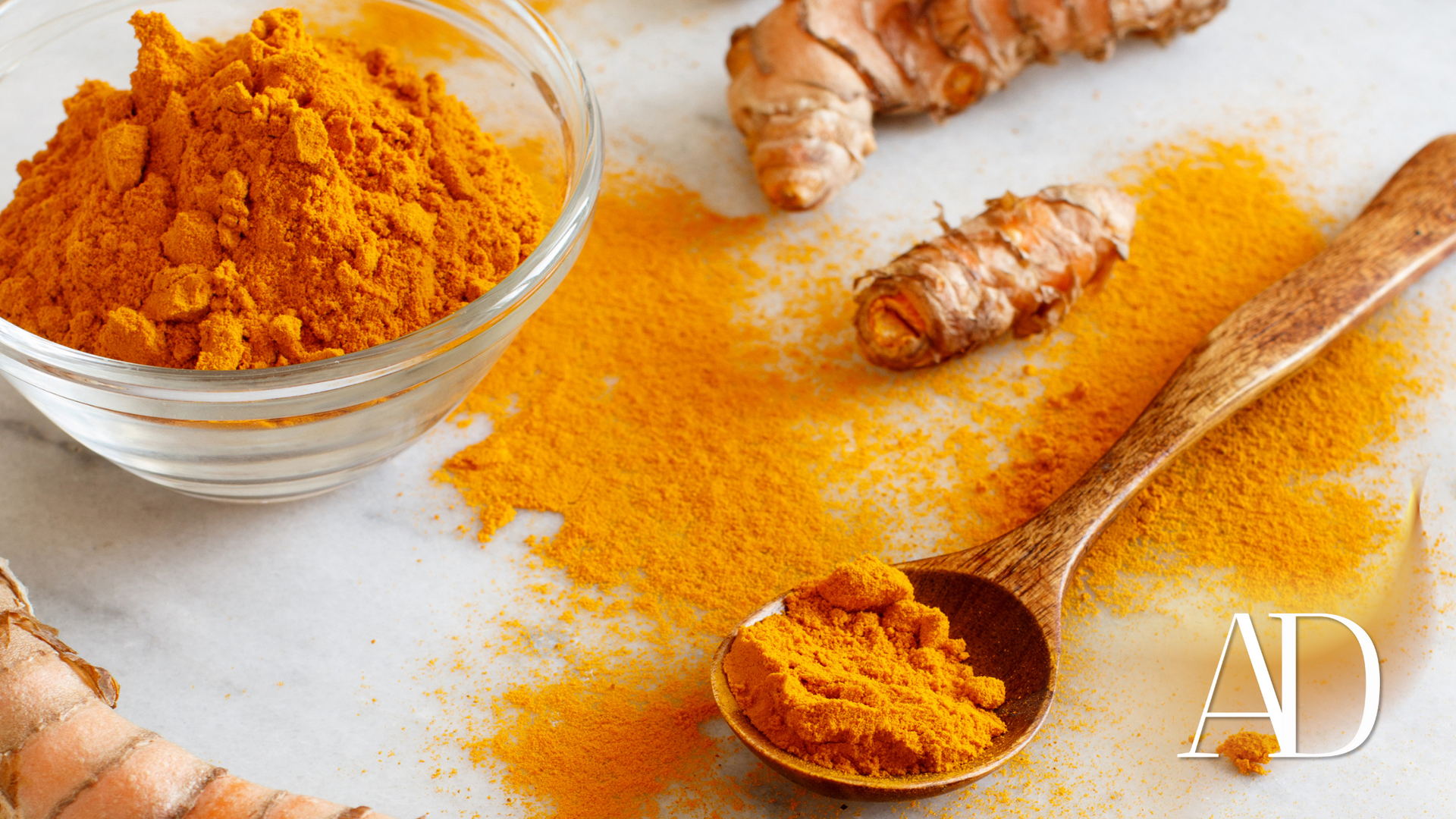 Turmeric powder in a glass bowl with roots and a wooden spoon on a white surface