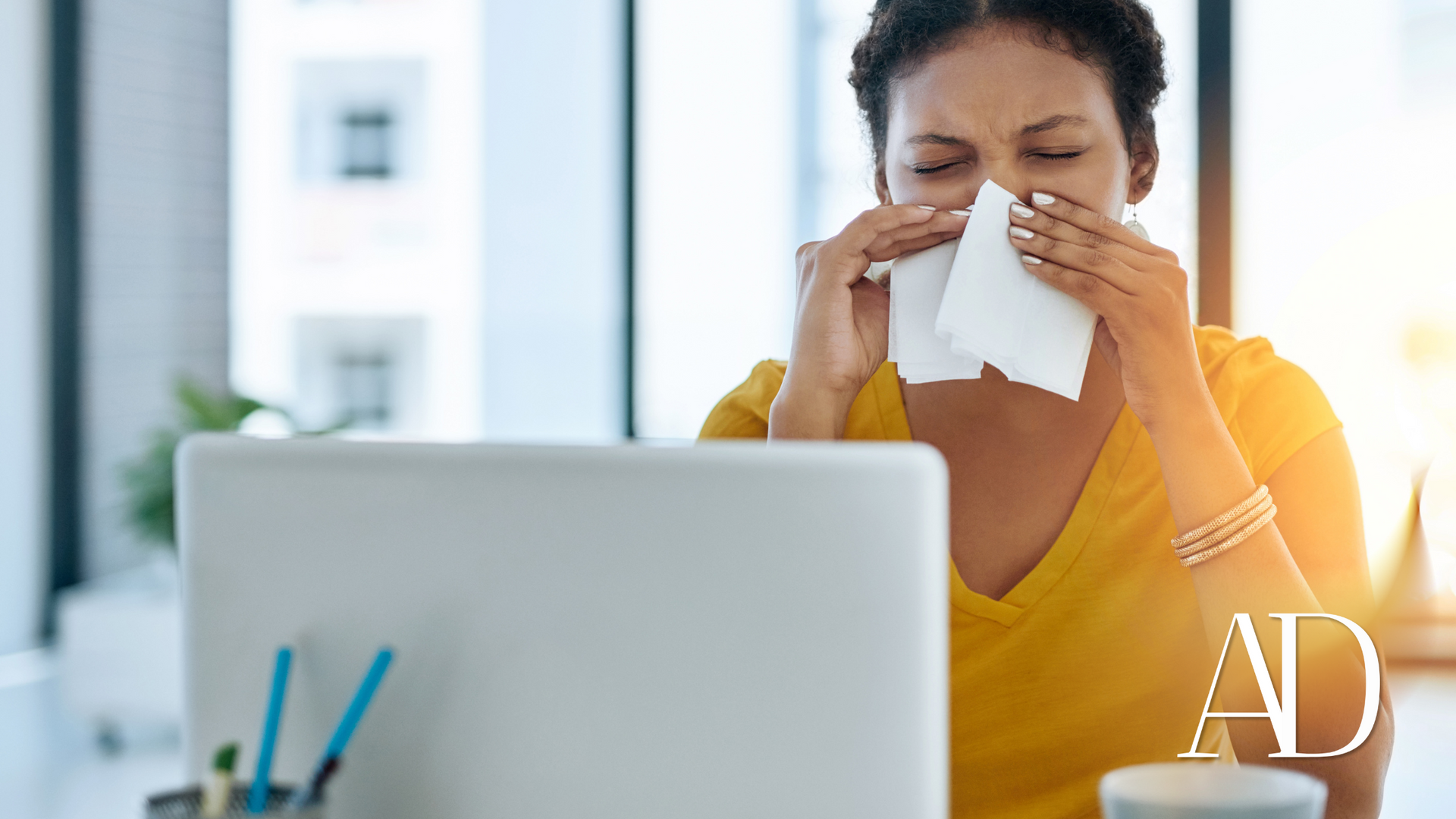 Person in a yellow sweater blowing their nose at a desk with a laptop and tissue box nearby