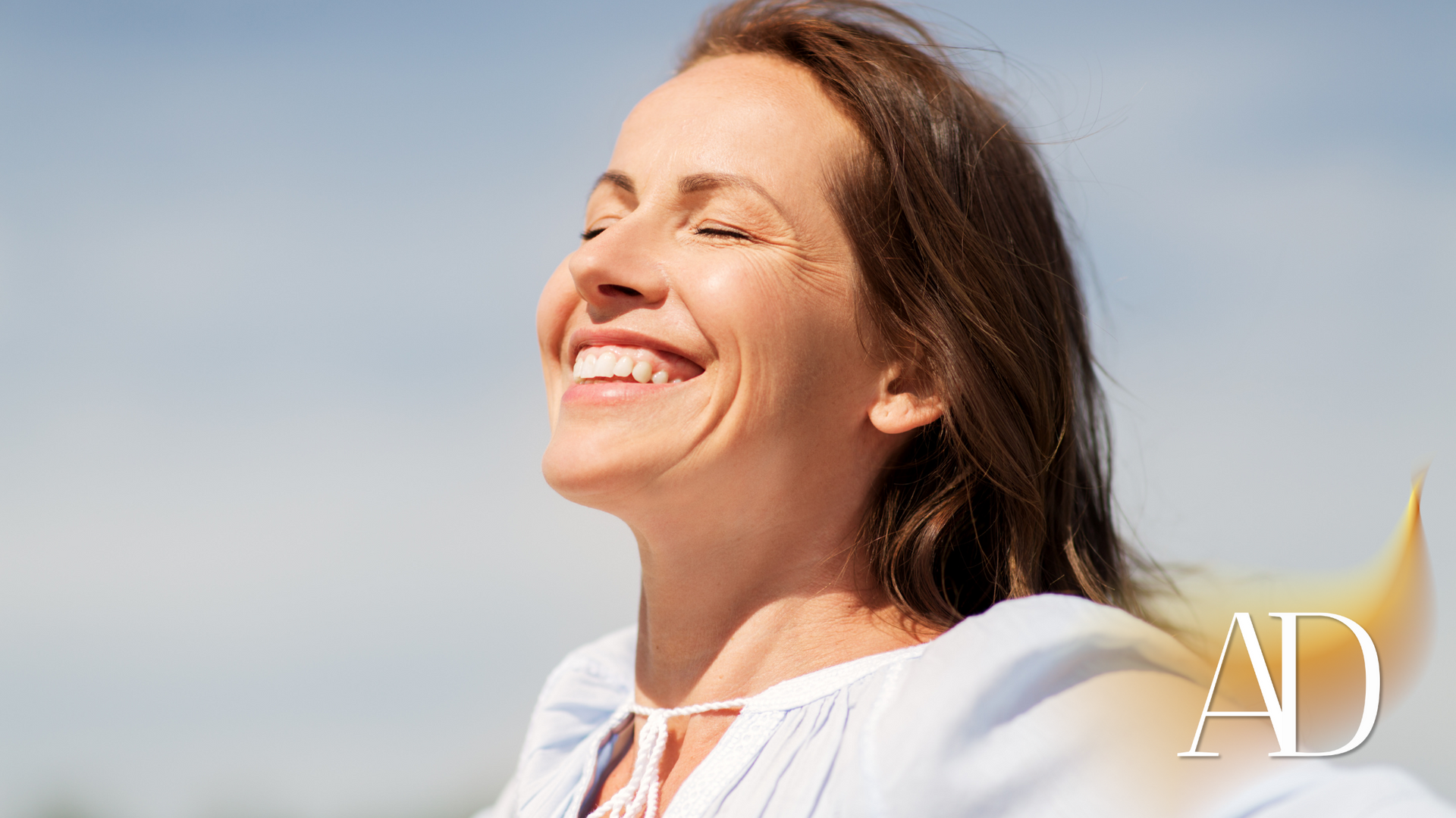 Smiling woman outdoors in bright sunlight, wearing a white top against a blue sky.