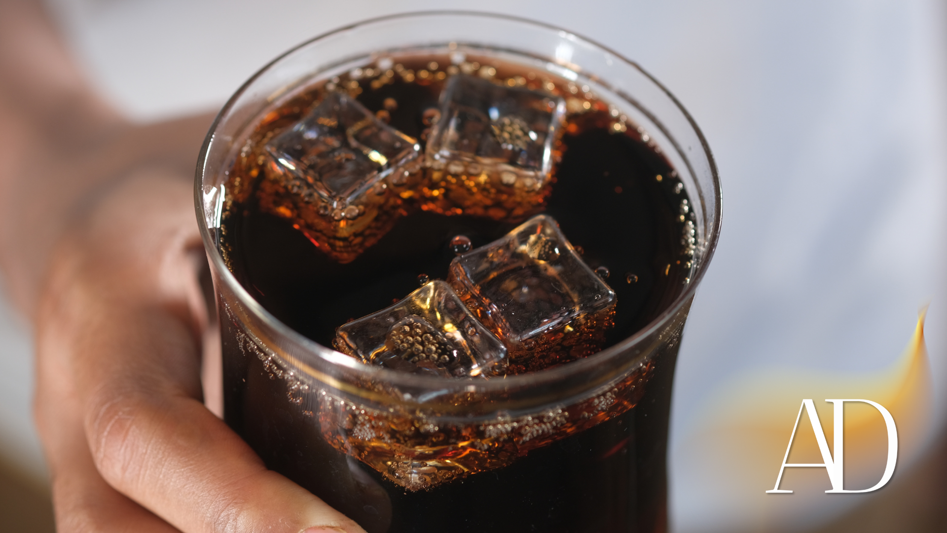 Close-up of a hand holding a glass of dark soda with ice cubes