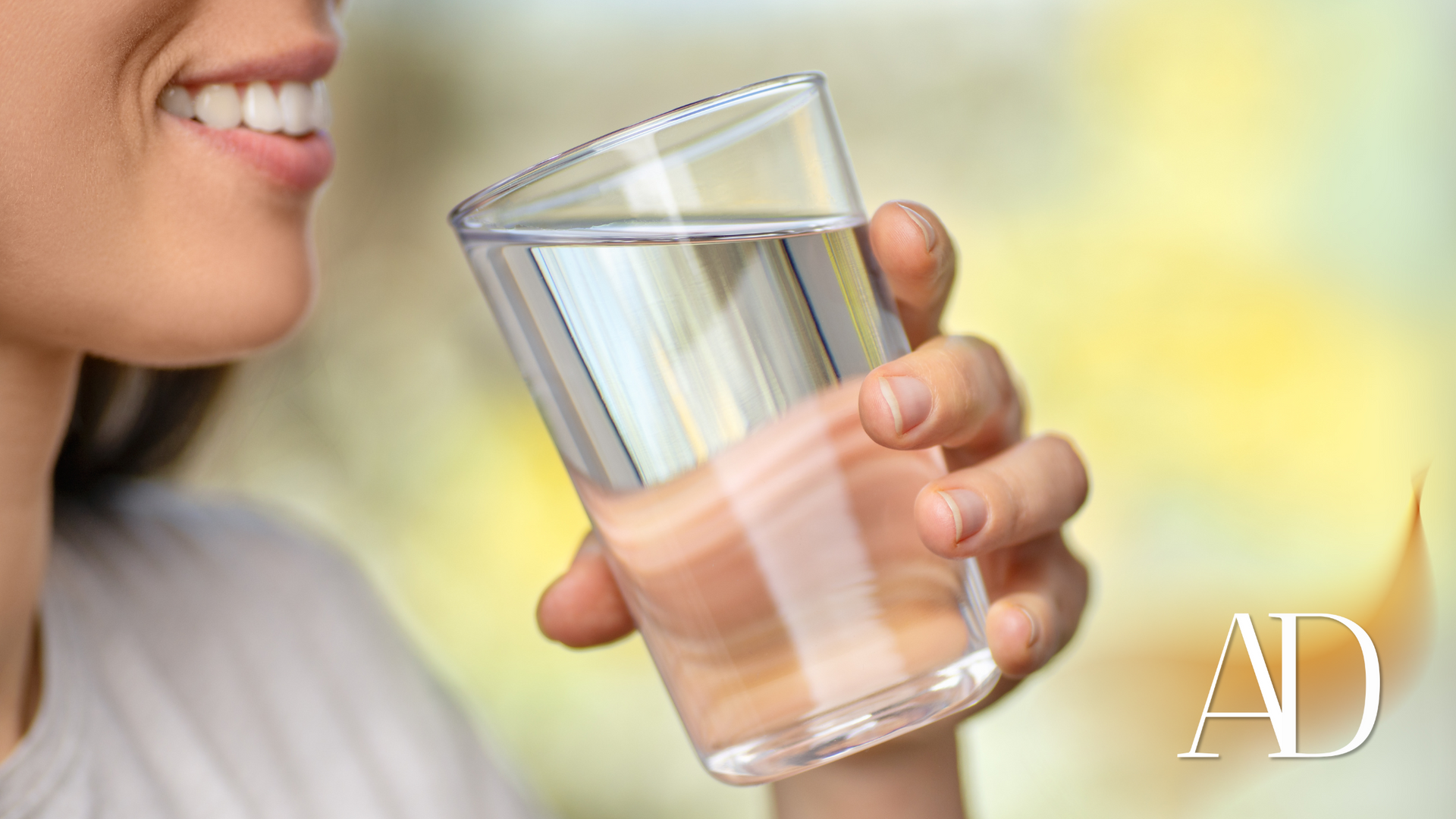 Person holding a glass of water near their mouth, smiling against a soft yellow background.