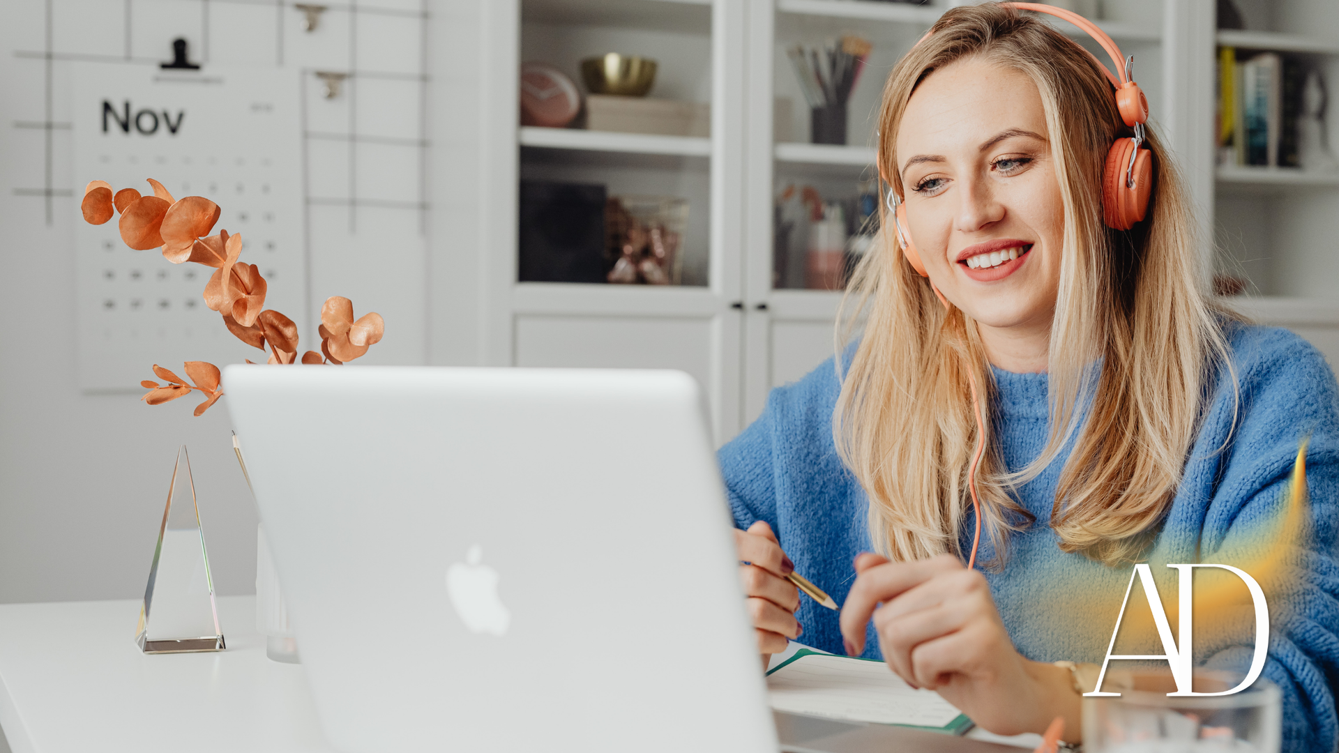 Woman in blue sweater smiling at laptop, holding a pen in a bright home office with flowers and shelves.