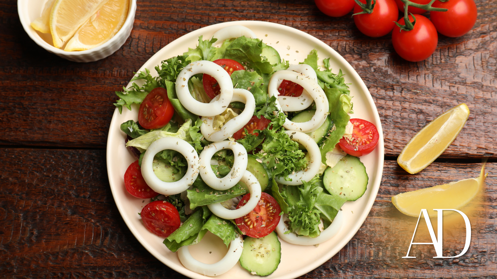 Fresh salad with cucumbers, tomatoes, and onion rings on a white plate with lemon wedges on a wooden table