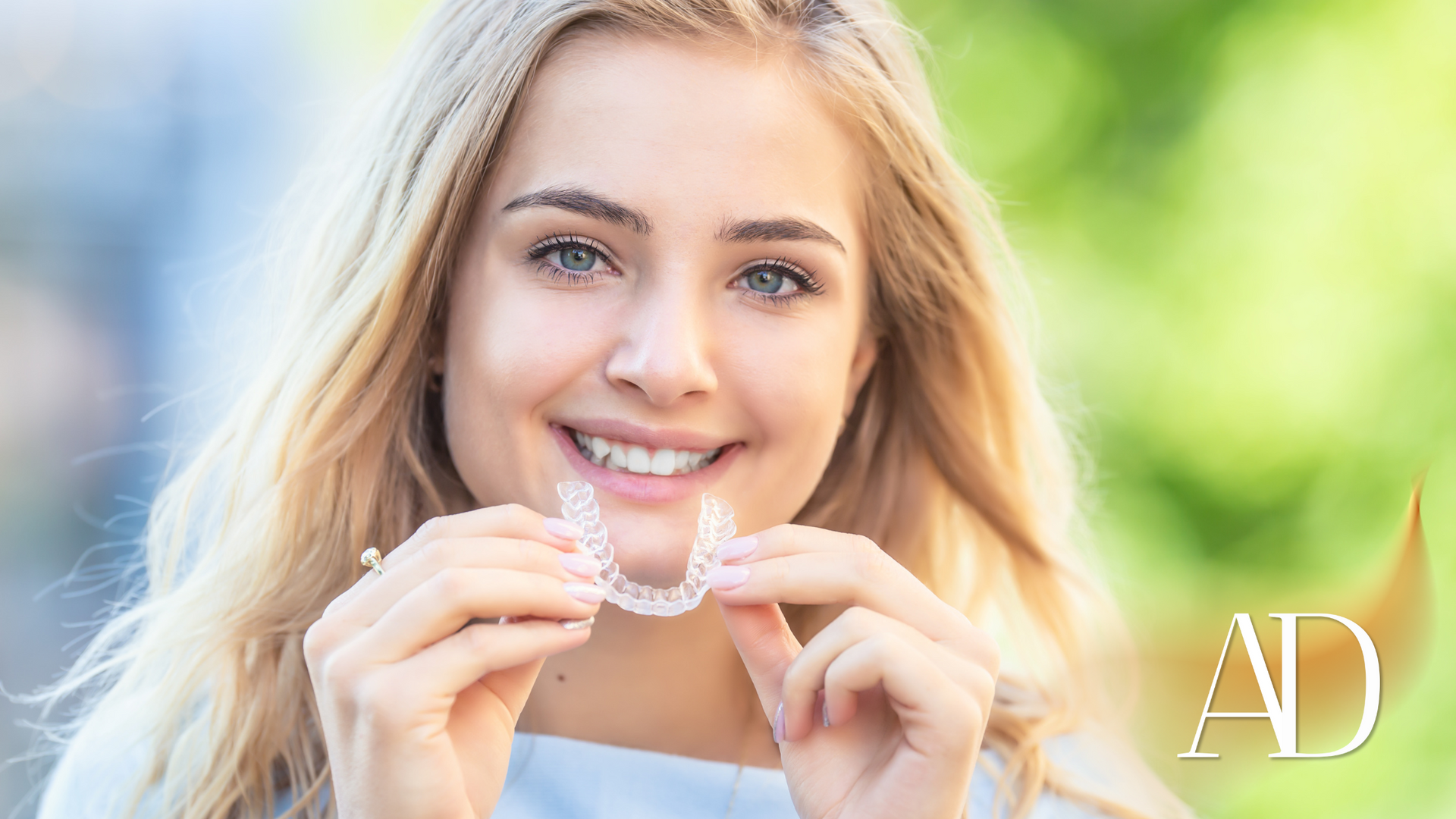 Smiling blonde woman holding clear aligners against a bright green outdoor background