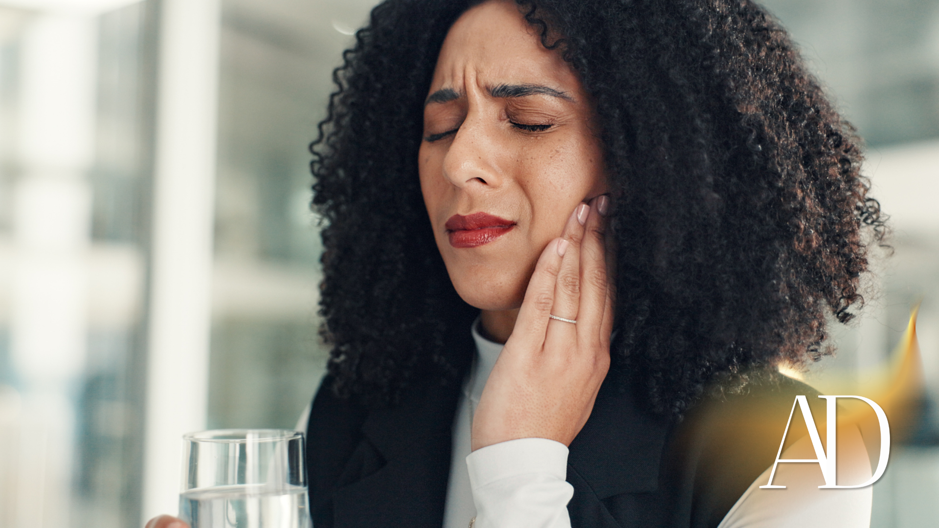 Woman holding her cheek with a pained expression beside a glass of water, suggesting toothache