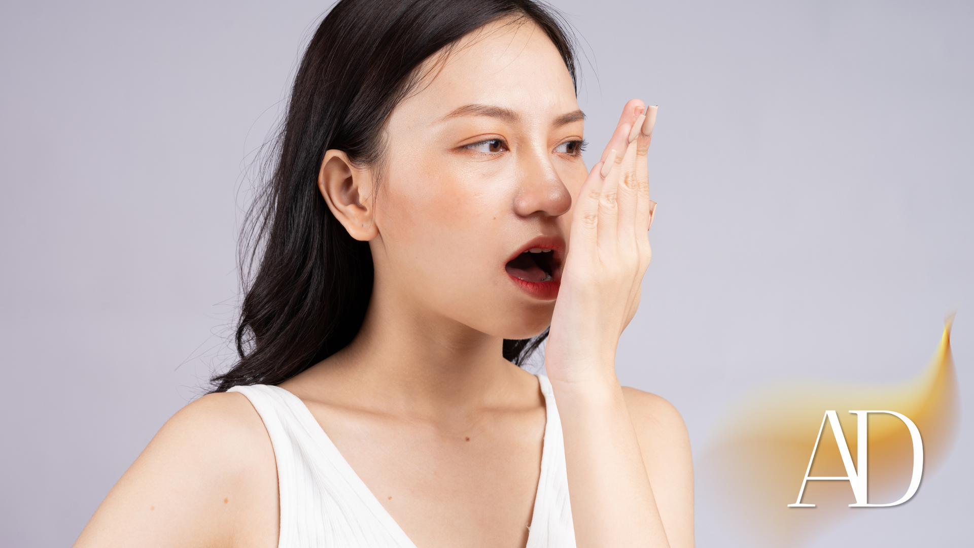 Woman in a white tank top covering her mouth with her hand, looking surprised against a light background.