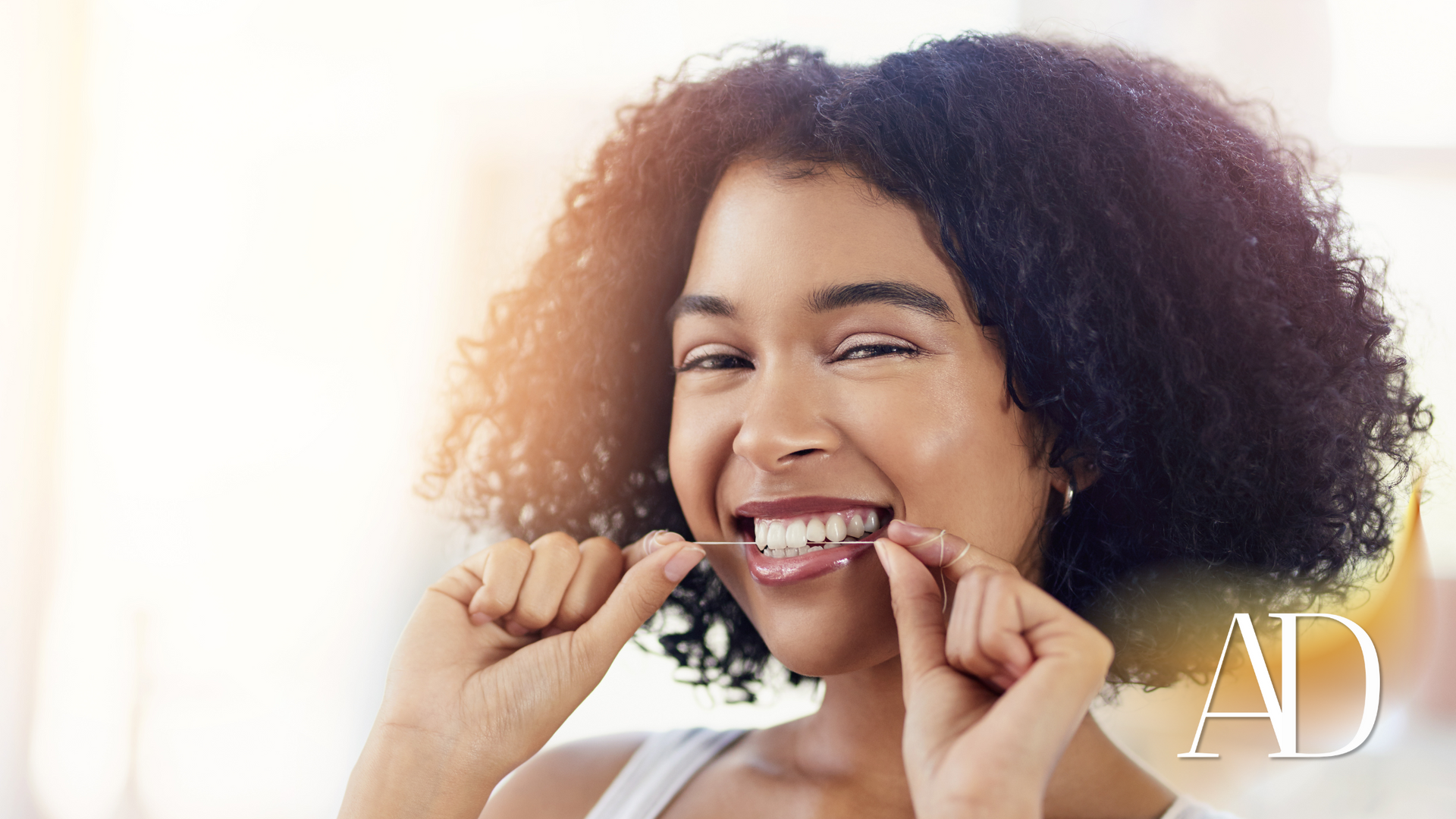 Smiling person flossing teeth in bright light, with curly hair and a white top.