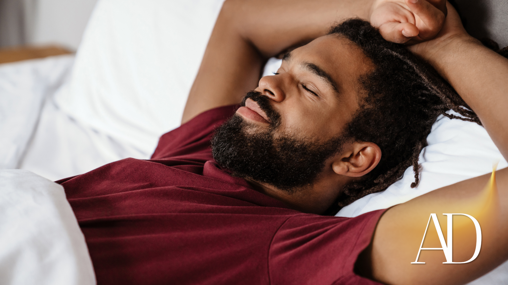Relaxing man lying on a bed with hands behind his head, wearing a maroon shirt, with an AD logo
