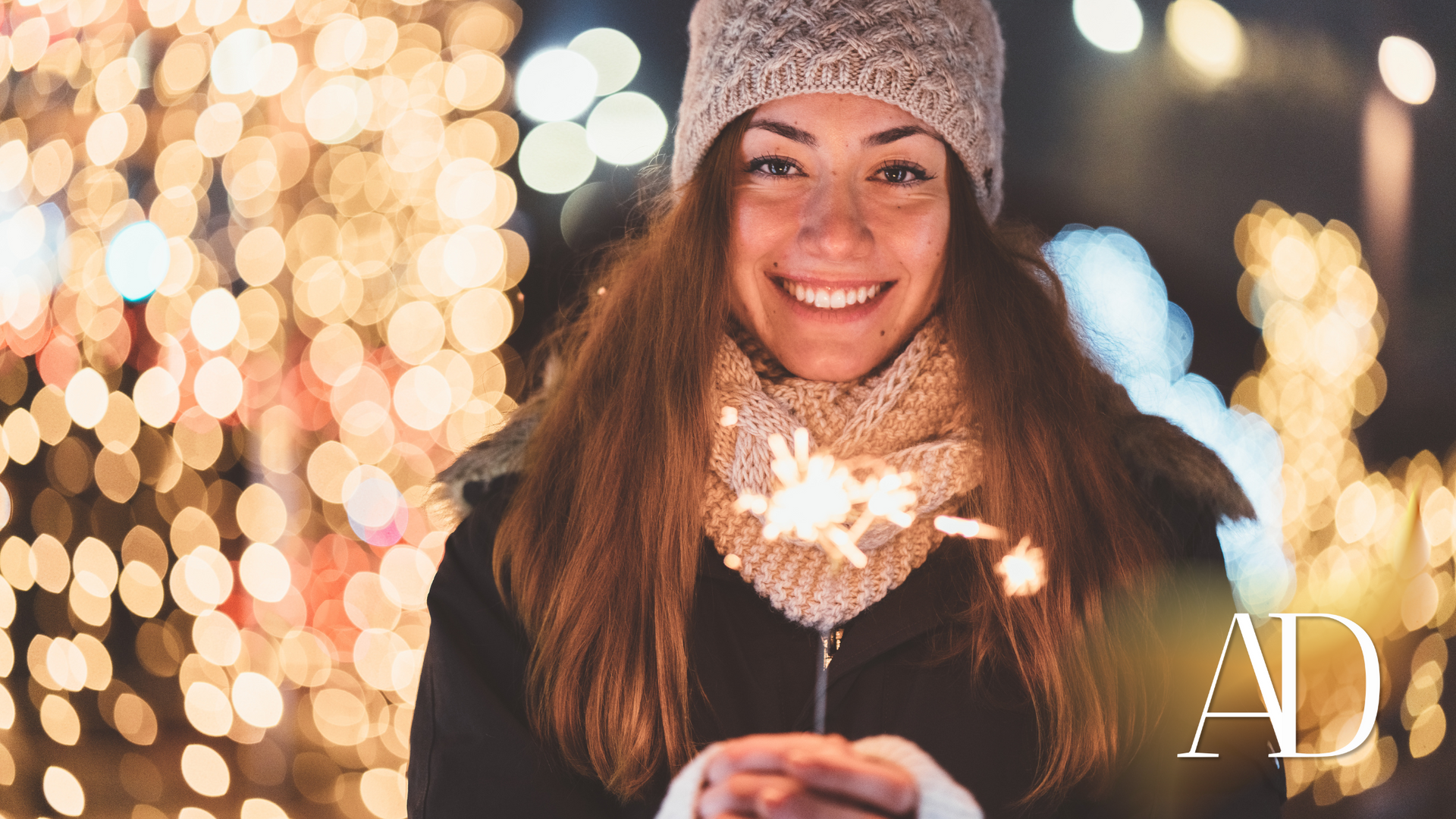 Smiling woman in a knit hat holding a lit sparkler with festive holiday lights behind her