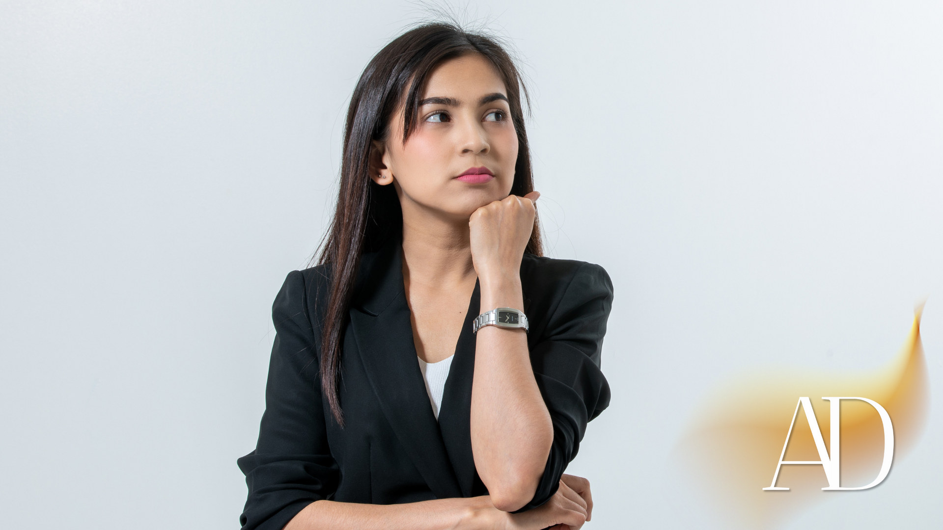 Woman in a black blazer resting her chin on her hand, looking thoughtful against a light background.
