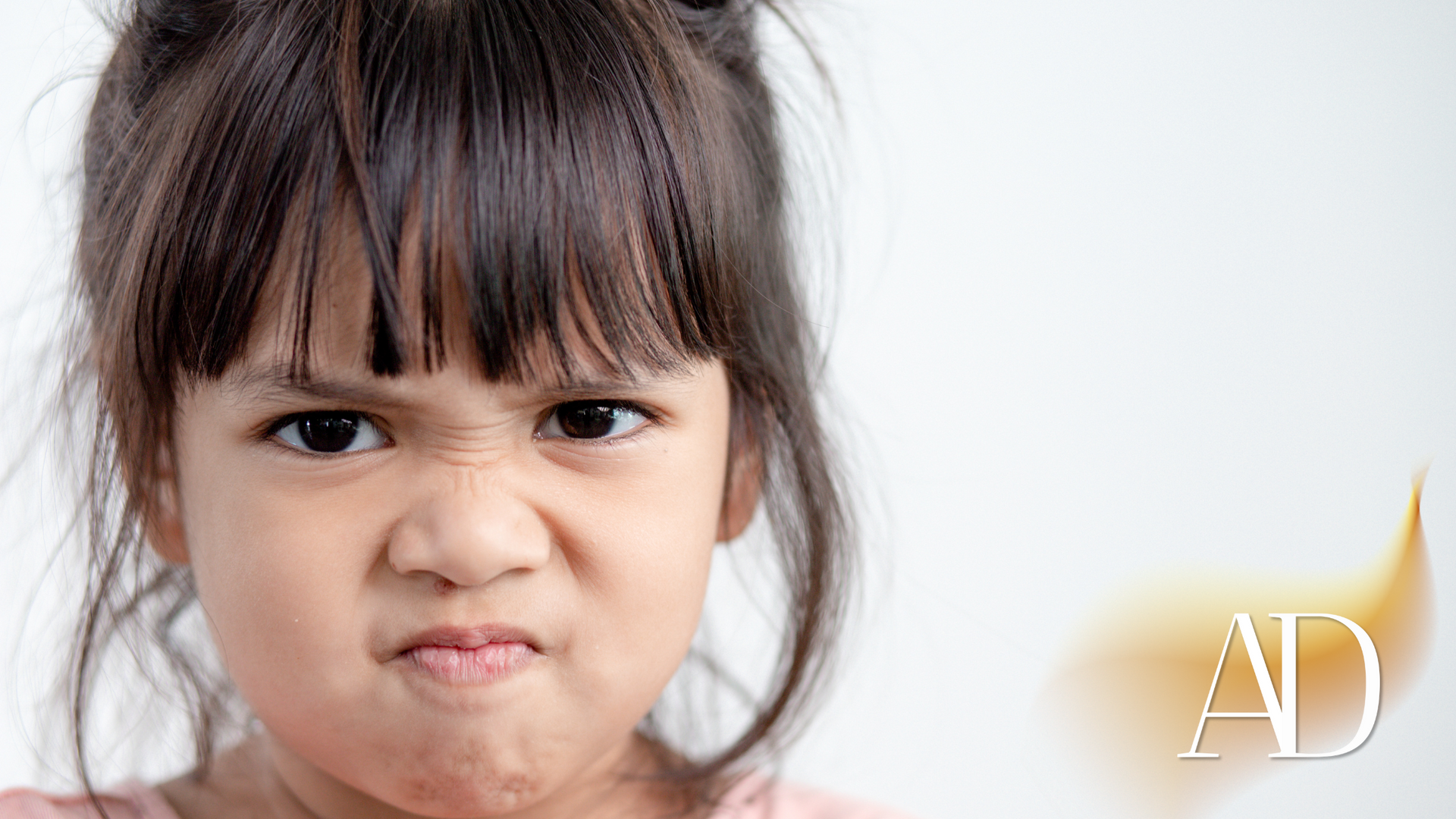 Child making a scrunched, angry face against a white background, with an “AD” watermark in the corner.