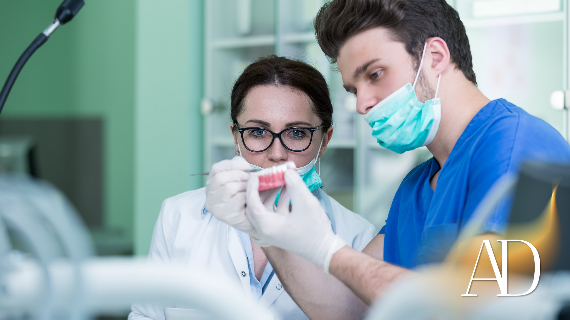 Dentist and assistant examining a dental model in a clinic, both wearing masks and gloves.