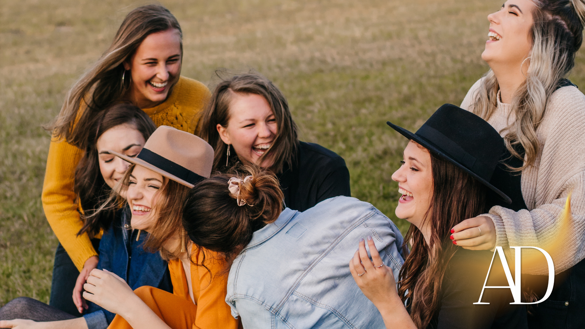 Friends laughing together outdoors on grass, with one person wearing a hat in the foreground