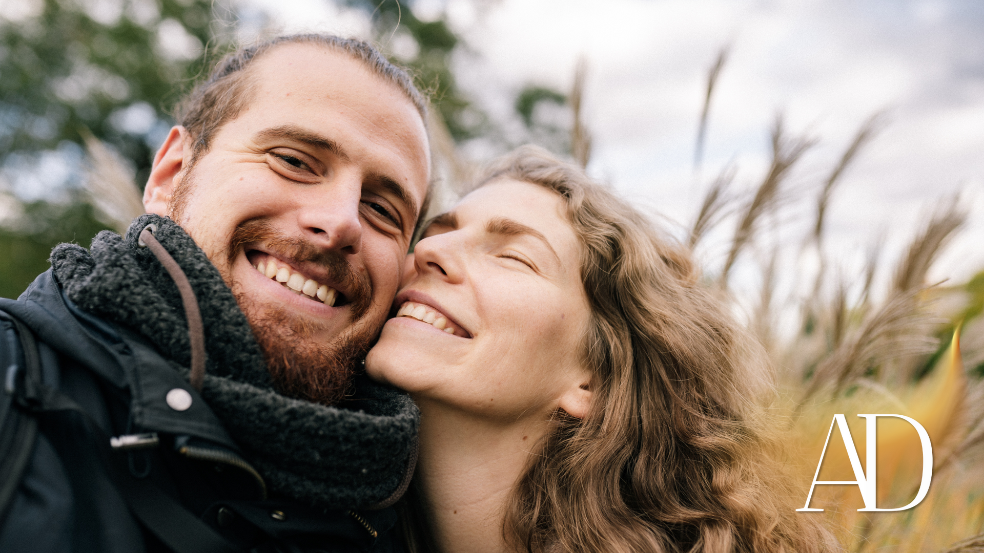 Smiling couple posing close together outdoors in a windy field, with a white “AD” logo in the corner.