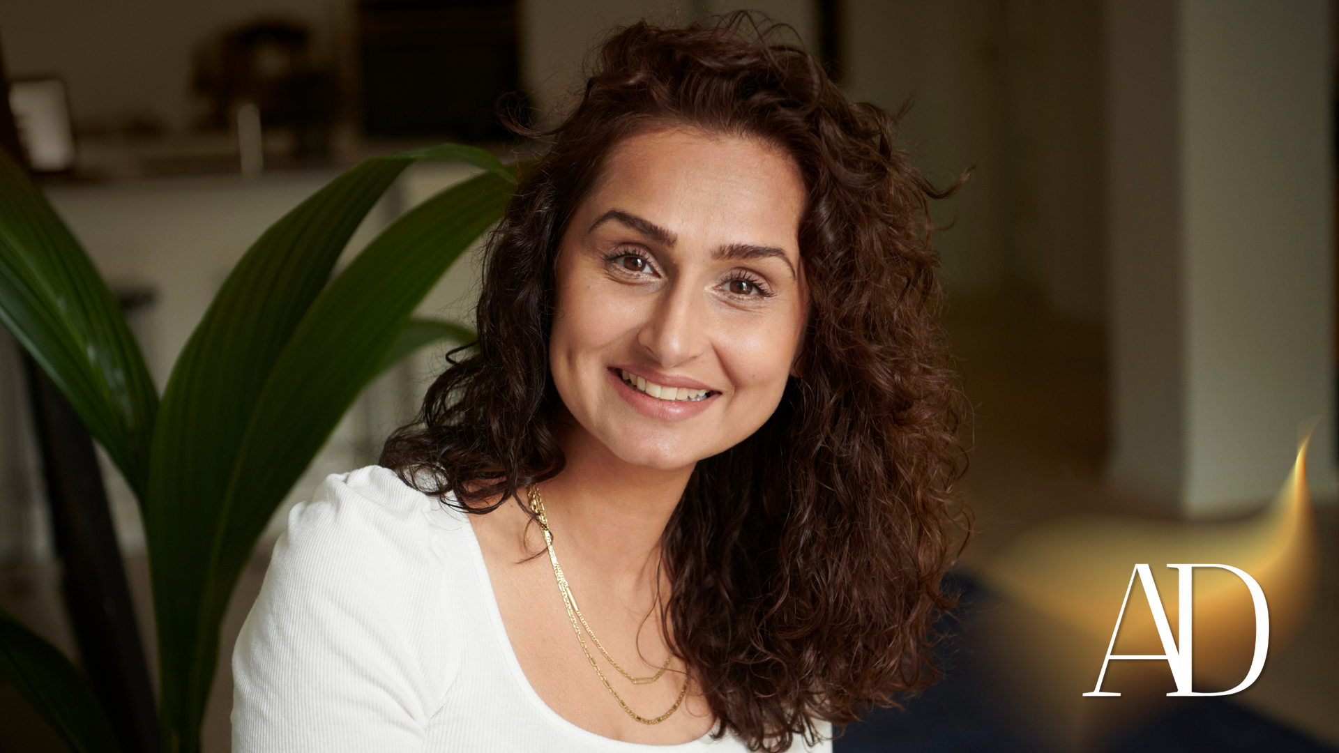 Smiling woman in a white top seated indoors, with a large green plant in the background.