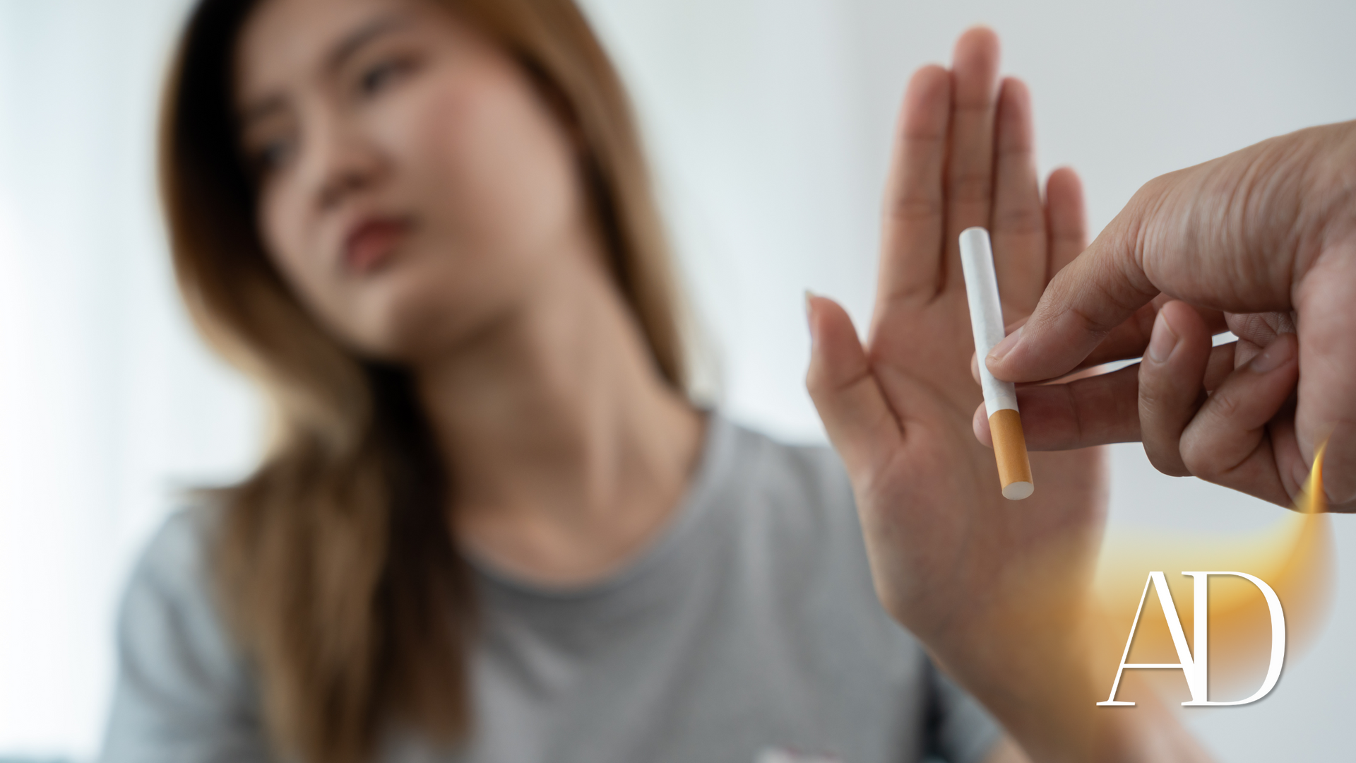 Cigarette held out toward a blurred person refusing it with a raised hand, indoors.