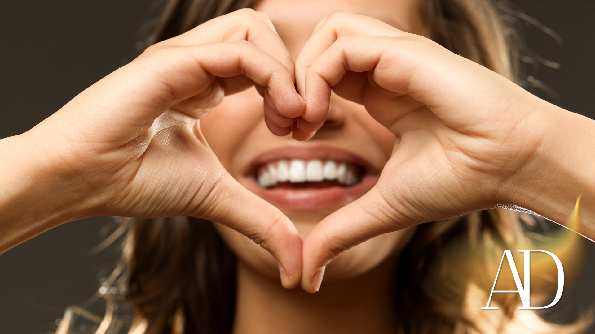 Hands forming a heart around a smiling mouth, with a small “AD” logo in the corner.